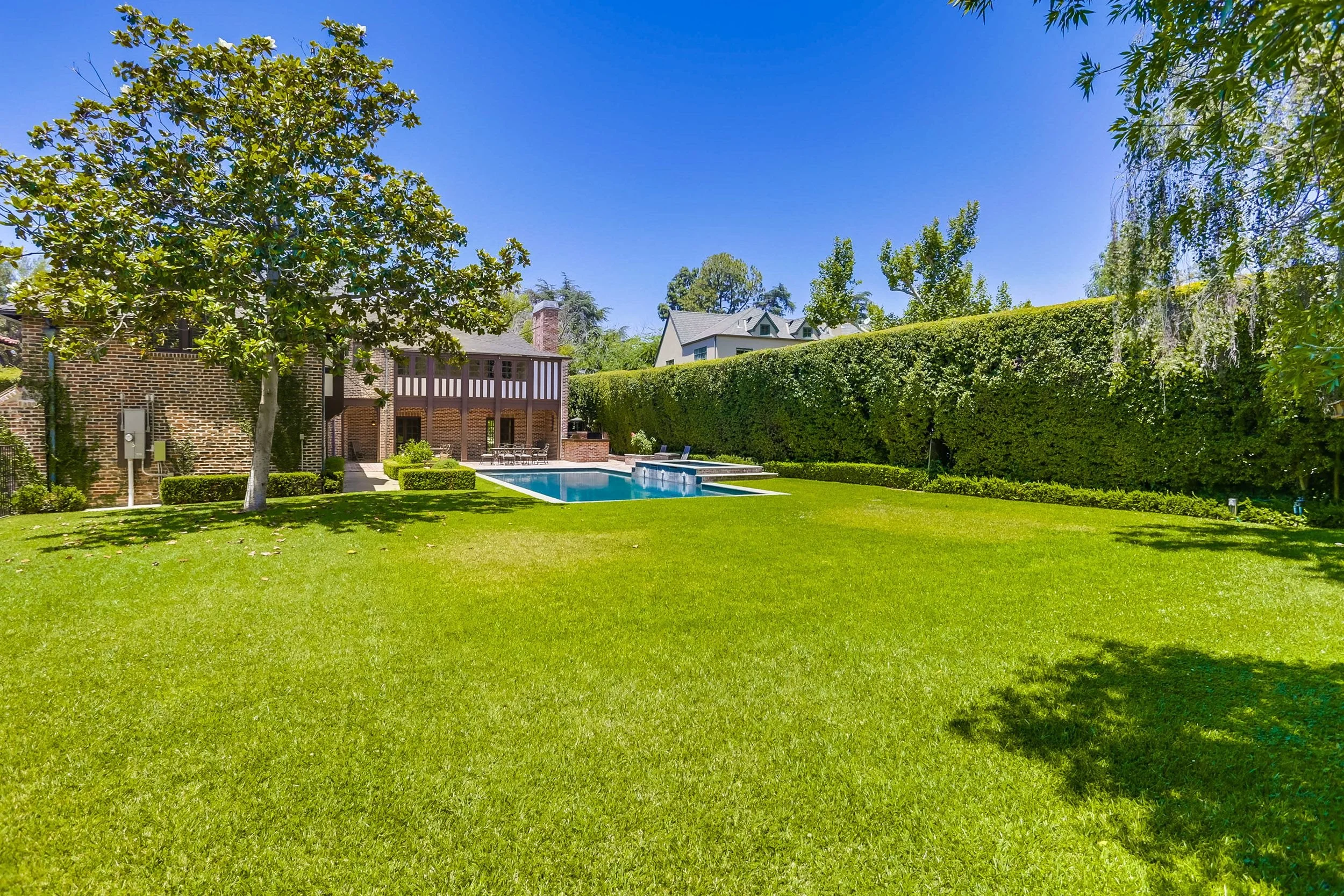 A backyard with a swimming pool, a well-maintained lawn, tall green hedges, and trees. A brick house with a second-story deck is visible in the background under a bright blue sky.