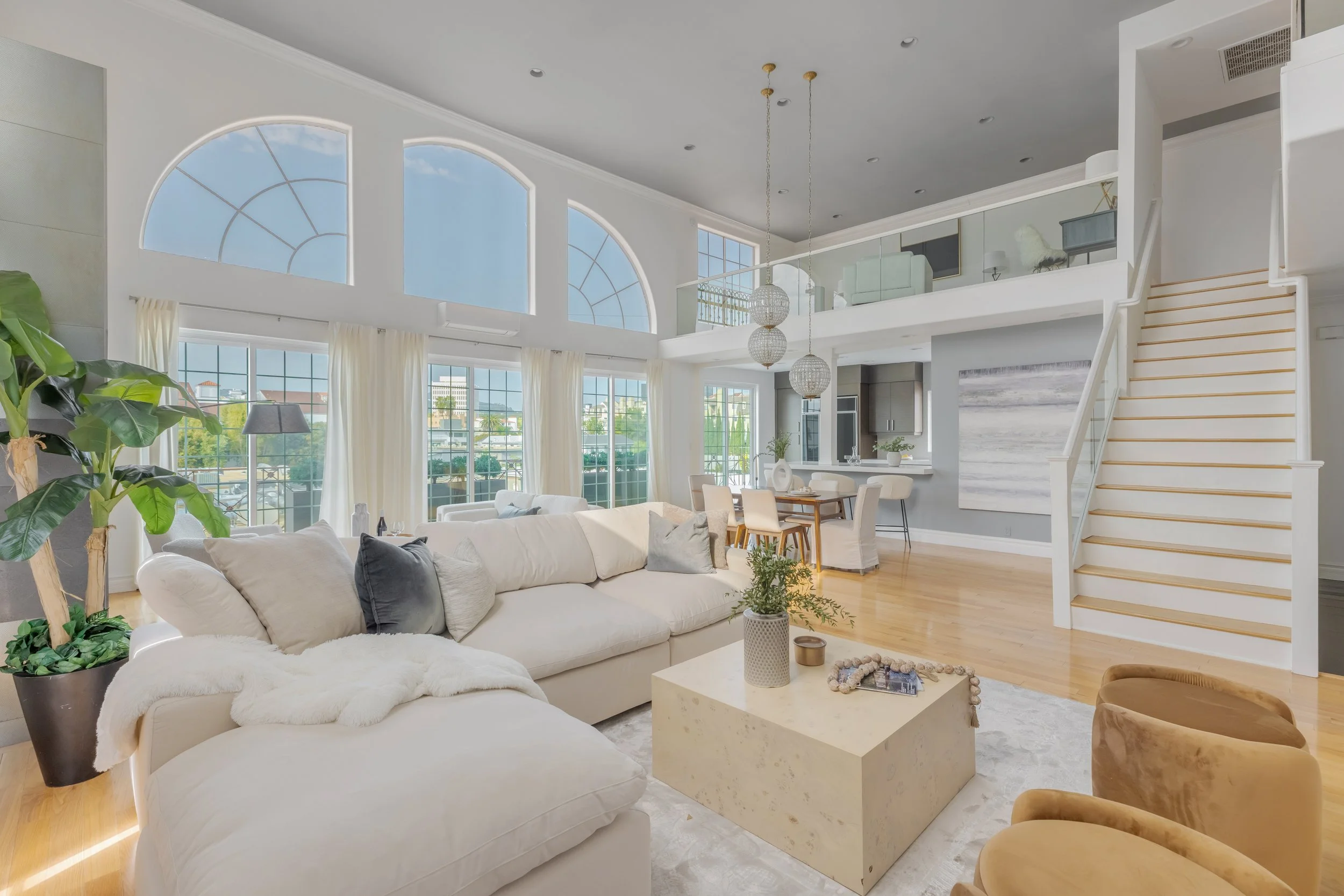 Bright living room with large windows, white sofas with gray pillows, a white marble coffee table, wooden flooring, a staircase with white railing, decorative pendant lights, and a view of the outdoors.