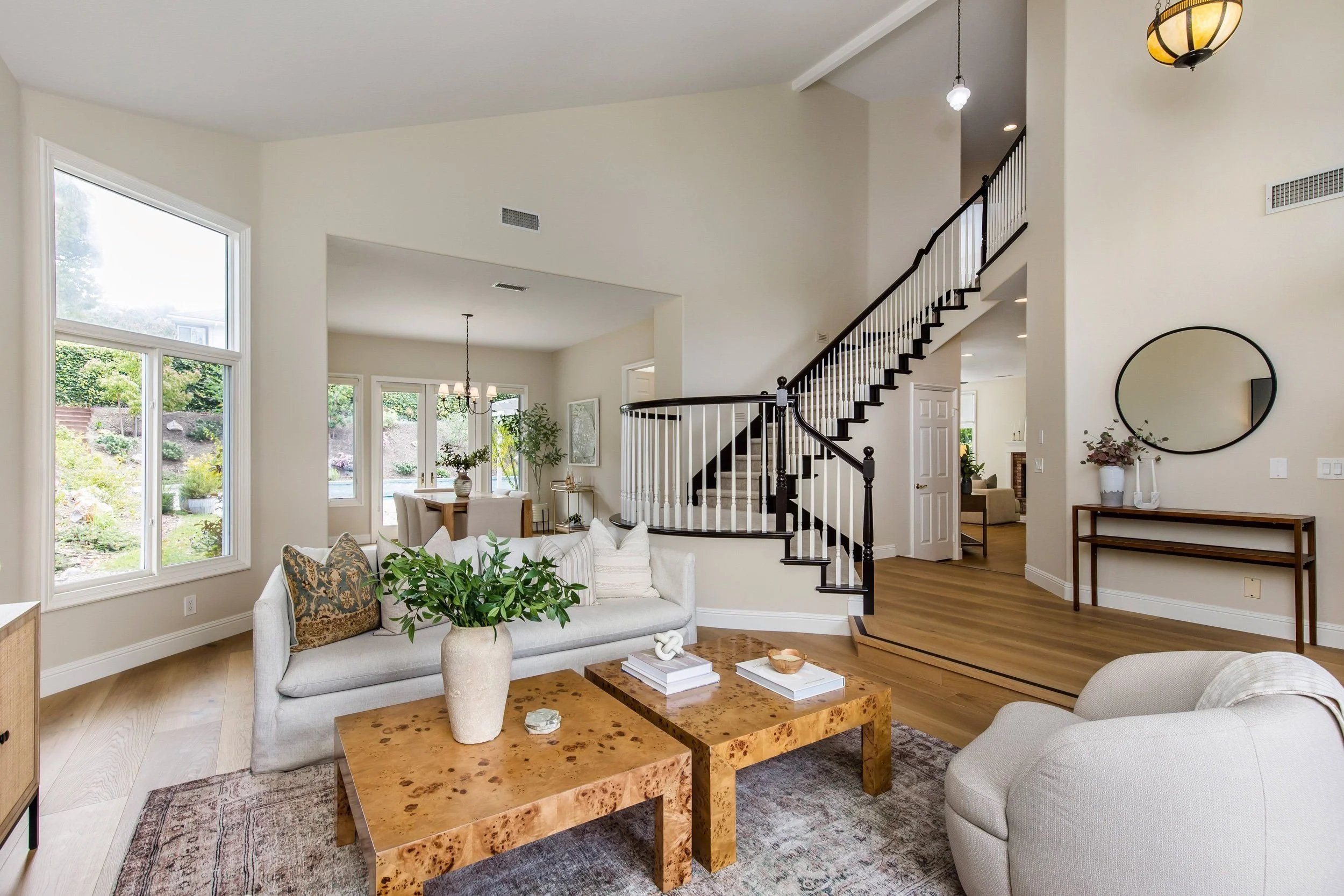 Bright living room with large windows, white sofas, a wooden coffee table, plants, and a staircase with black handrail leading upstairs.
