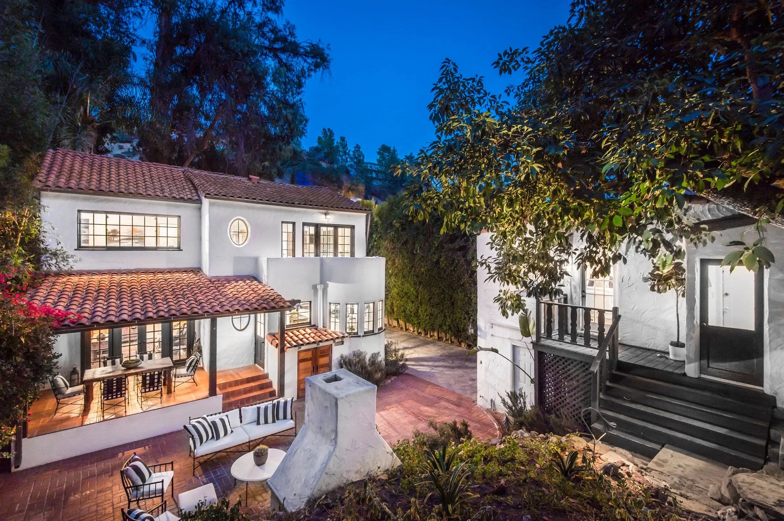 A view of a multi-story house with white walls, tiled red roof, and large windows, surrounded by trees and outdoor furniture, taken during early evening or dusk.