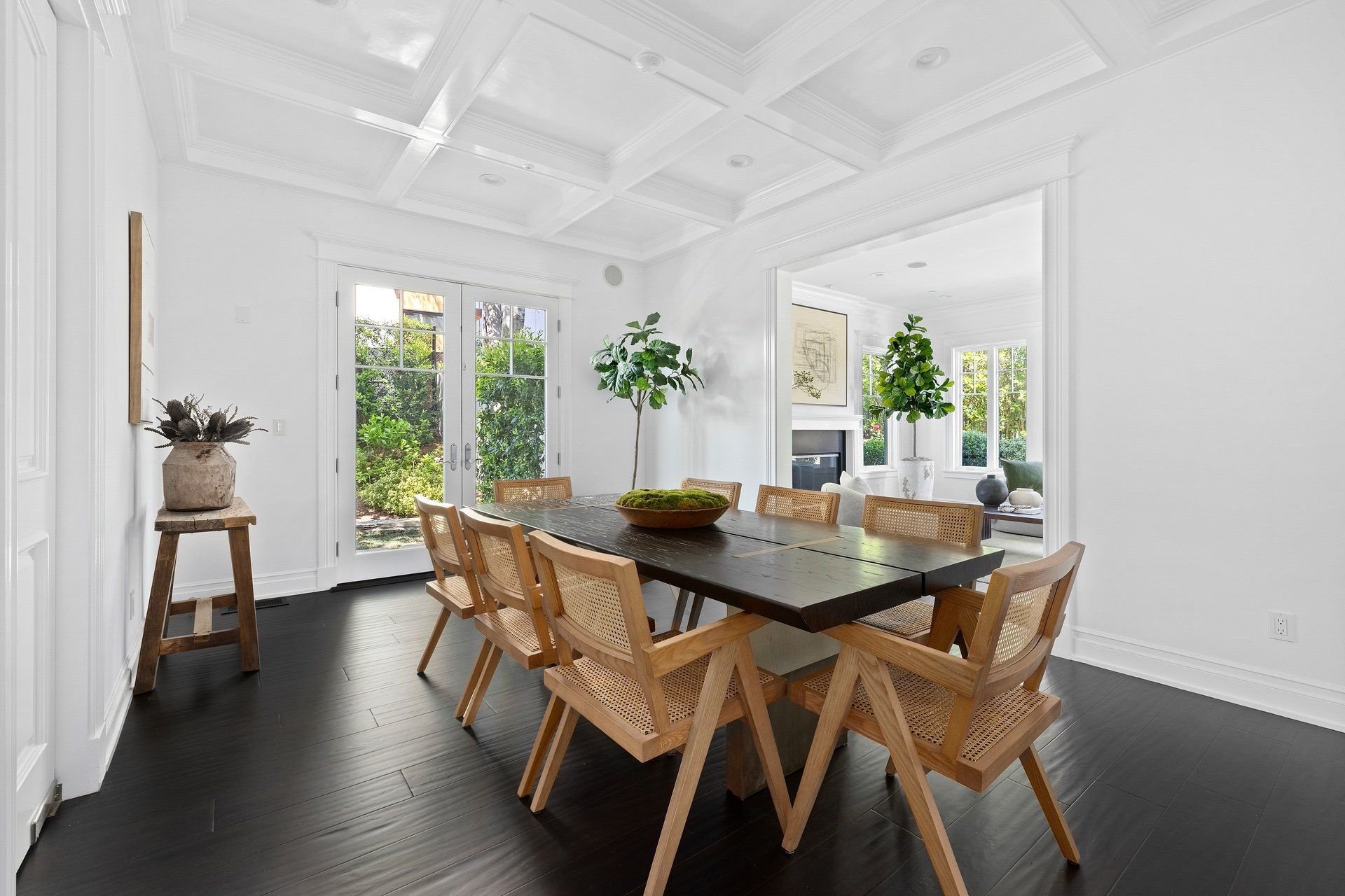 Dining room with a dark wood table, six wooden chairs with woven seats, and a large windowed door leading outside. The room has white walls, a coffered ceiling, and decorative plants.