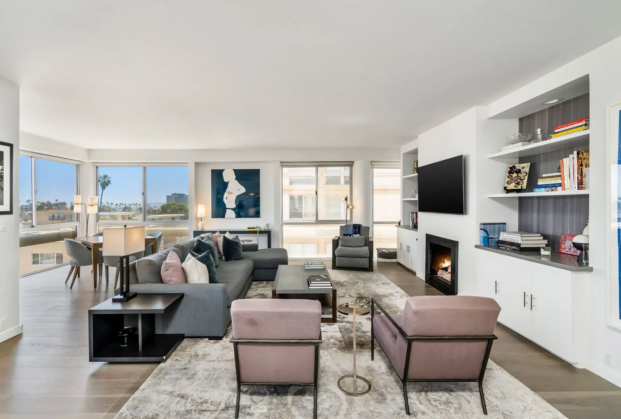Modern living room with gray sectional sofa, pink armchair, and a fireplace, large windows with city views, and wall art.