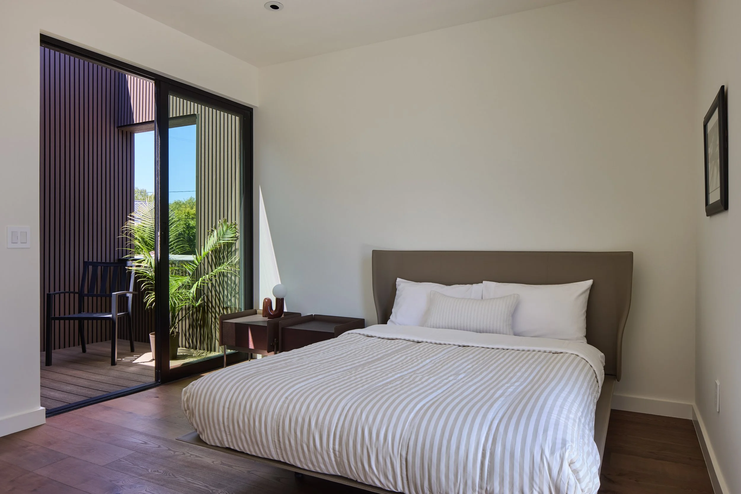 A modern bedroom with a bed, nightstand, and sliding glass door leading to a balcony with outdoor seating and plants, overlooking greenery.