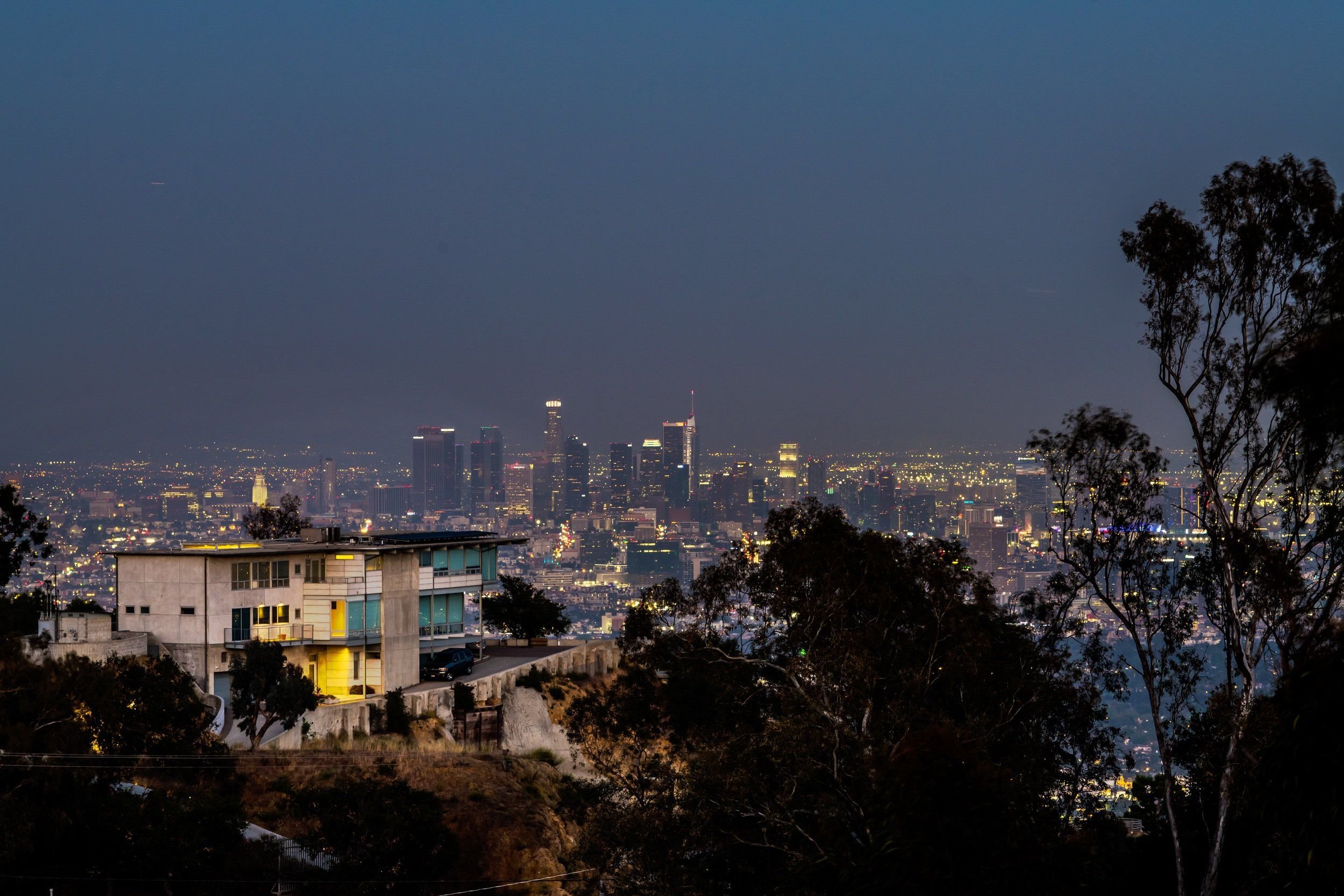 Nighttime cityscape of Los Angeles with tall buildings illuminated, view from a hillside with trees and residential buildings in the foreground.