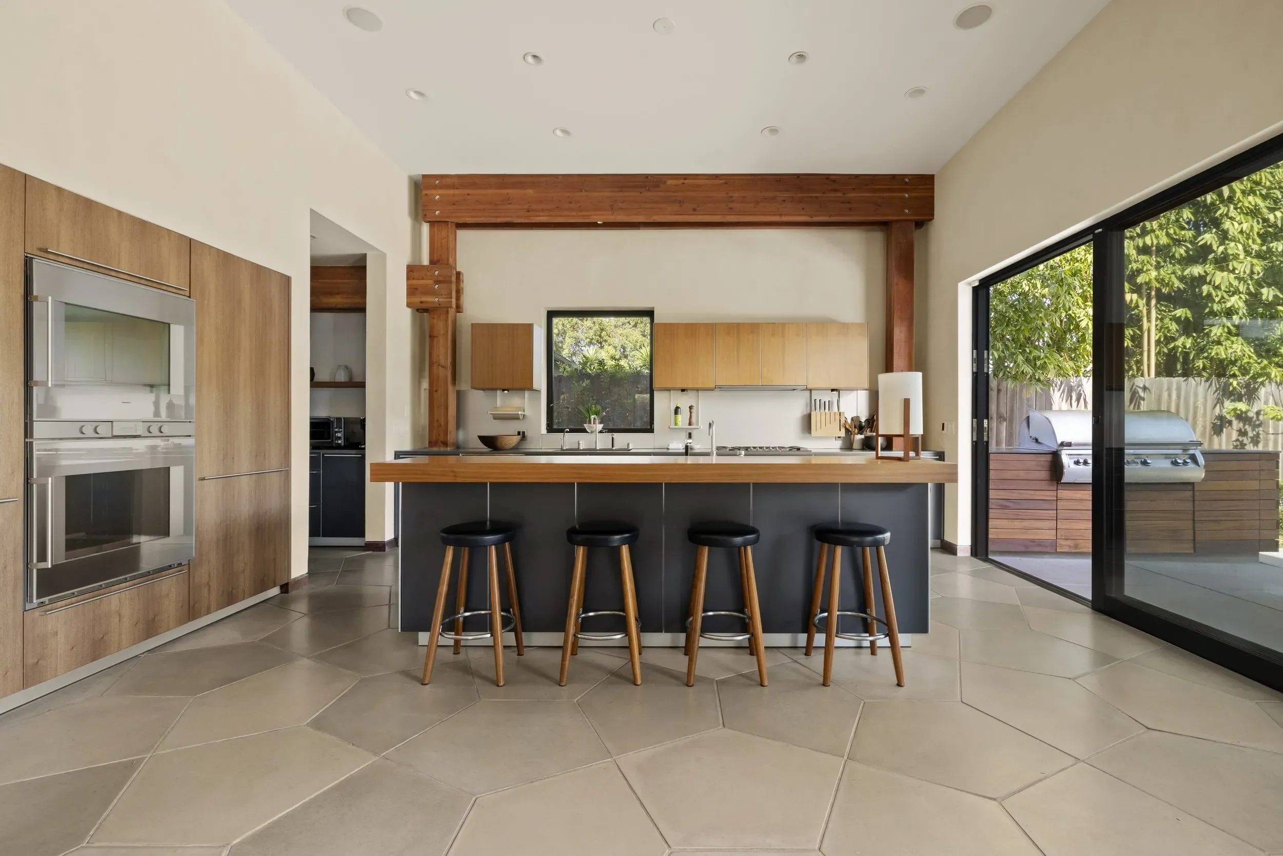 Modern kitchen with wood cabinets, a large island with black stools, stainless steel appliances, a window, and sliding glass doors leading to an outdoor barbecue area.