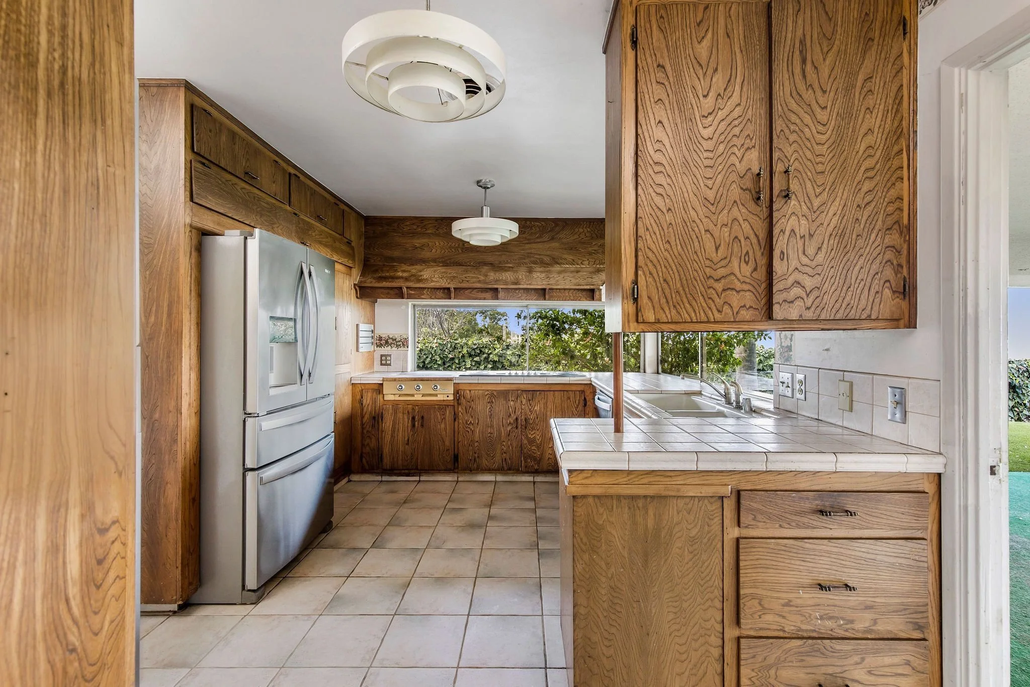 Kitchen with wooden cabinets, white tiled countertops, and a stainless steel refrigerator. Large window showing greenery outside, with ceiling lights and a tiled floor.