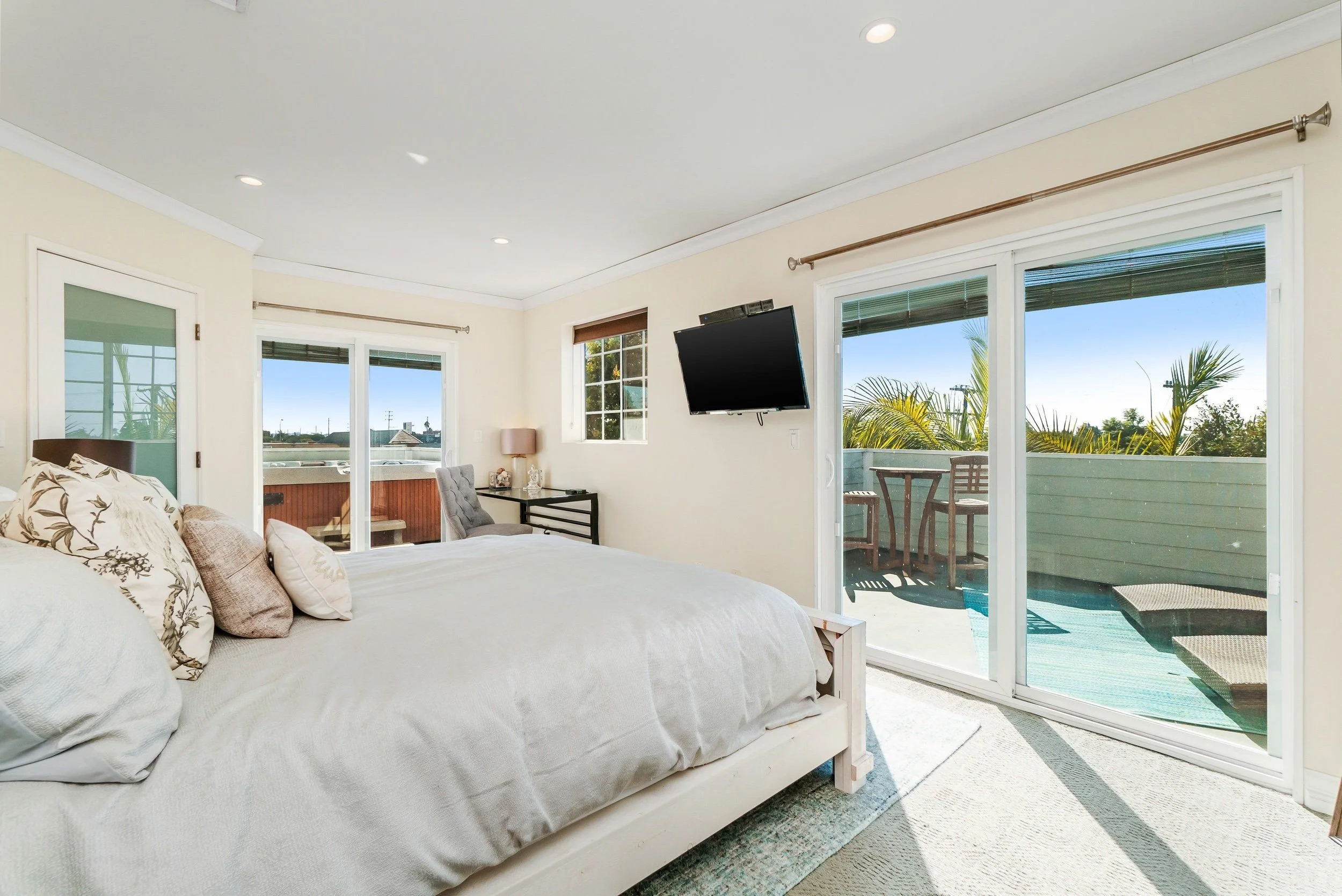 Bright bedroom with sliding glass doors leading to a balcony, beige walls, a bed with white and floral pillows, a desk with a chair, and a wall-mounted flat-screen TV.