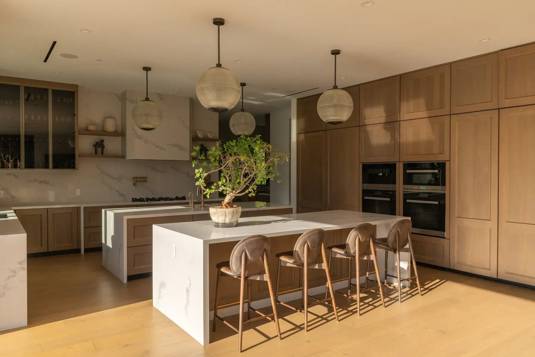 Modern kitchen with wooden cabinets, white marble island, and a potted plant on the island. Pendant lights hang from the ceiling, and built-in ovens are integrated into the cabinetry.