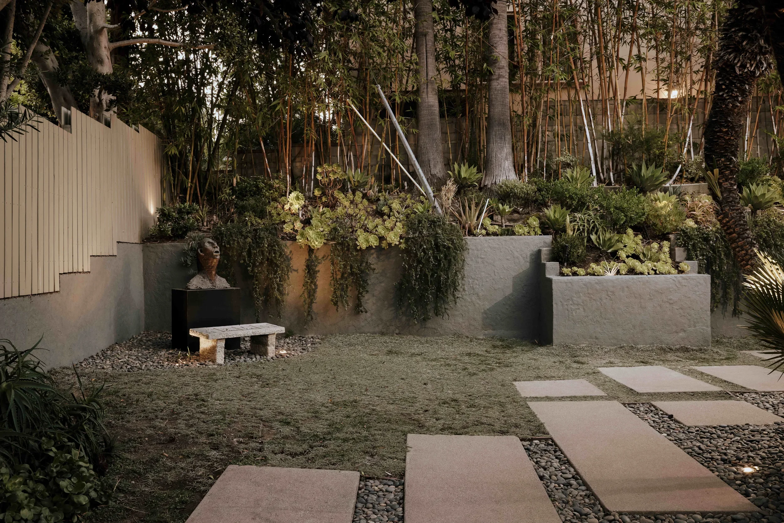 A backyard garden scene at dusk with a concrete bench, stone pathway, lush green plants, succulents, a beige fence, a stone statue with a head and shoulders, and large trees with palm trees in the background.