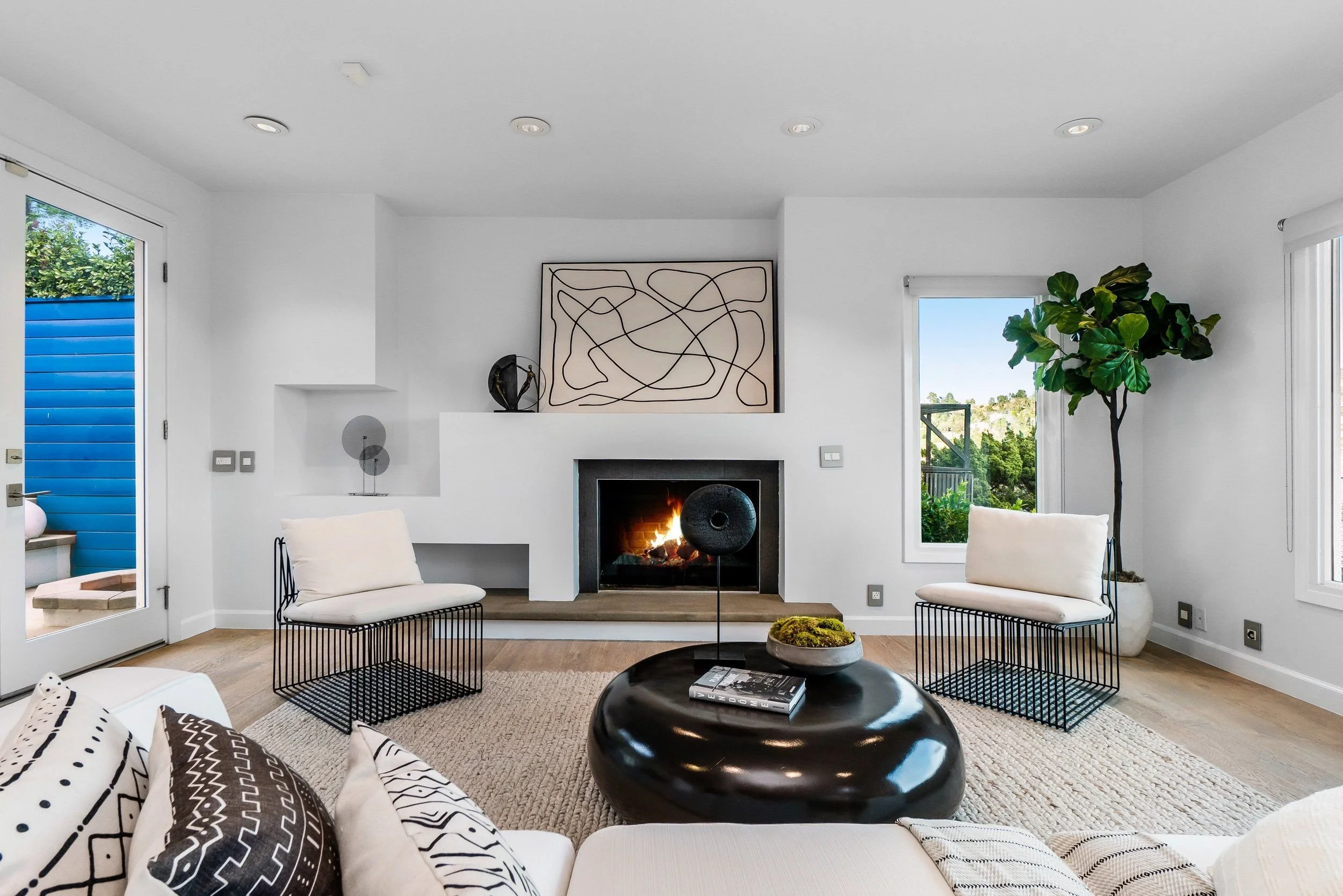 Modern living room with white walls, a fireplace, abstract artwork, large windows showing greenery outside, and contemporary furniture including white chairs, black wireframe tables, and patterned pillows.