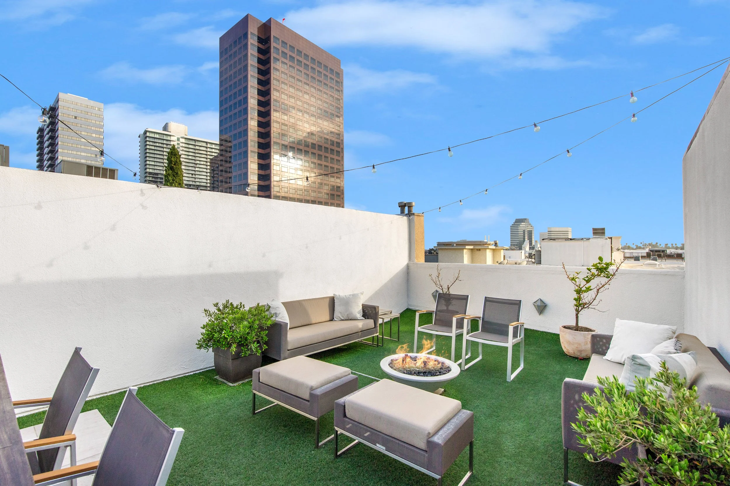 Rooftop terrace with outdoor seating, a fire pit, greenery, string lights, and city skyscrapers in the background on a clear day.