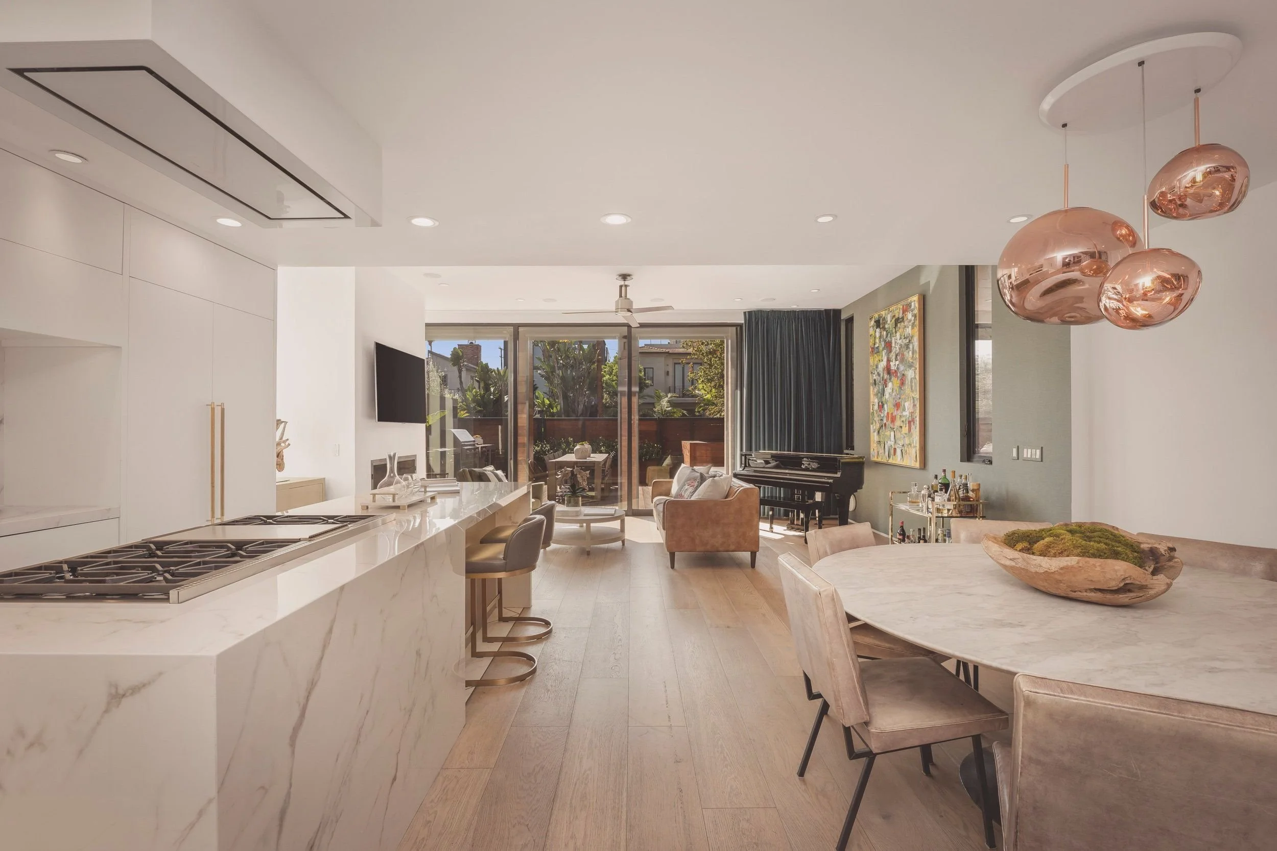 Open-concept living and kitchen area with white marble island, beige chairs, a black piano, and a large window with curtains revealing outdoor greenery.