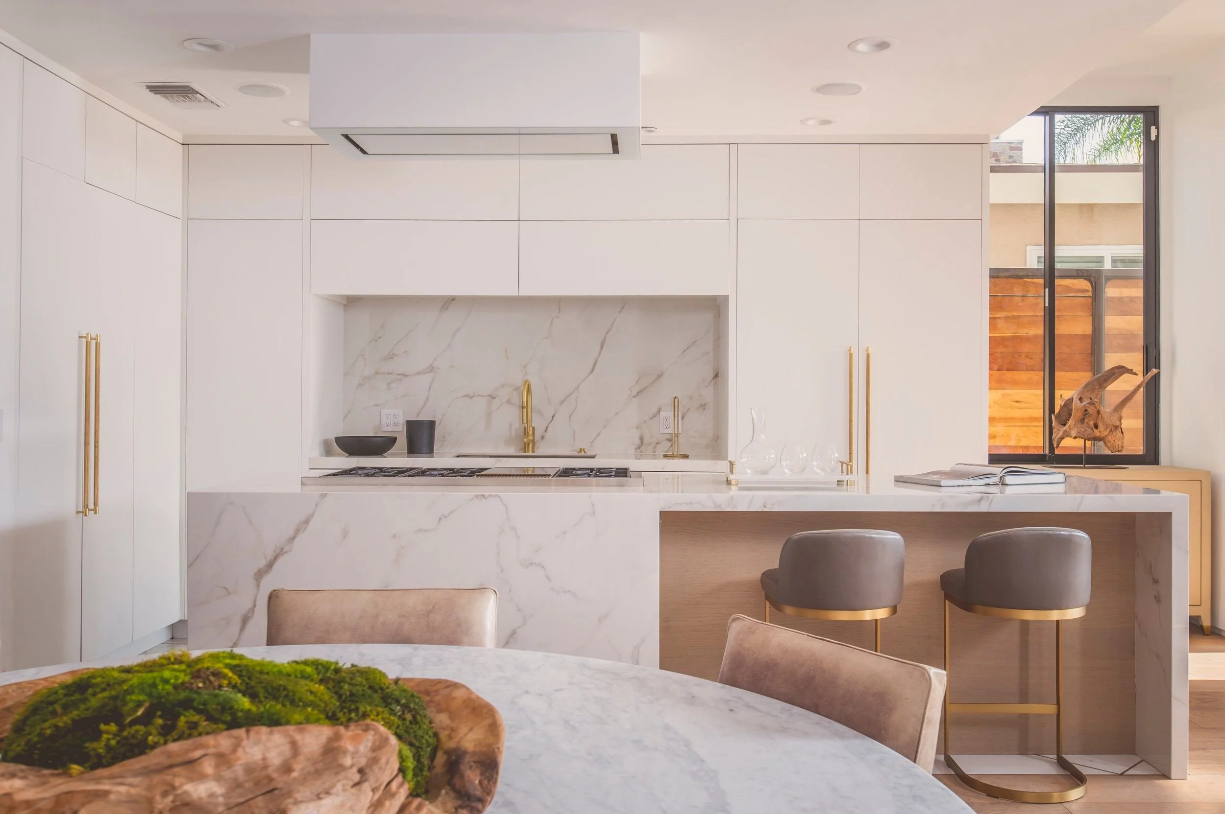 Modern kitchen with white cabinetry, marble countertops, a gold faucet, and a window with a view of a wooden fence and palm trees.