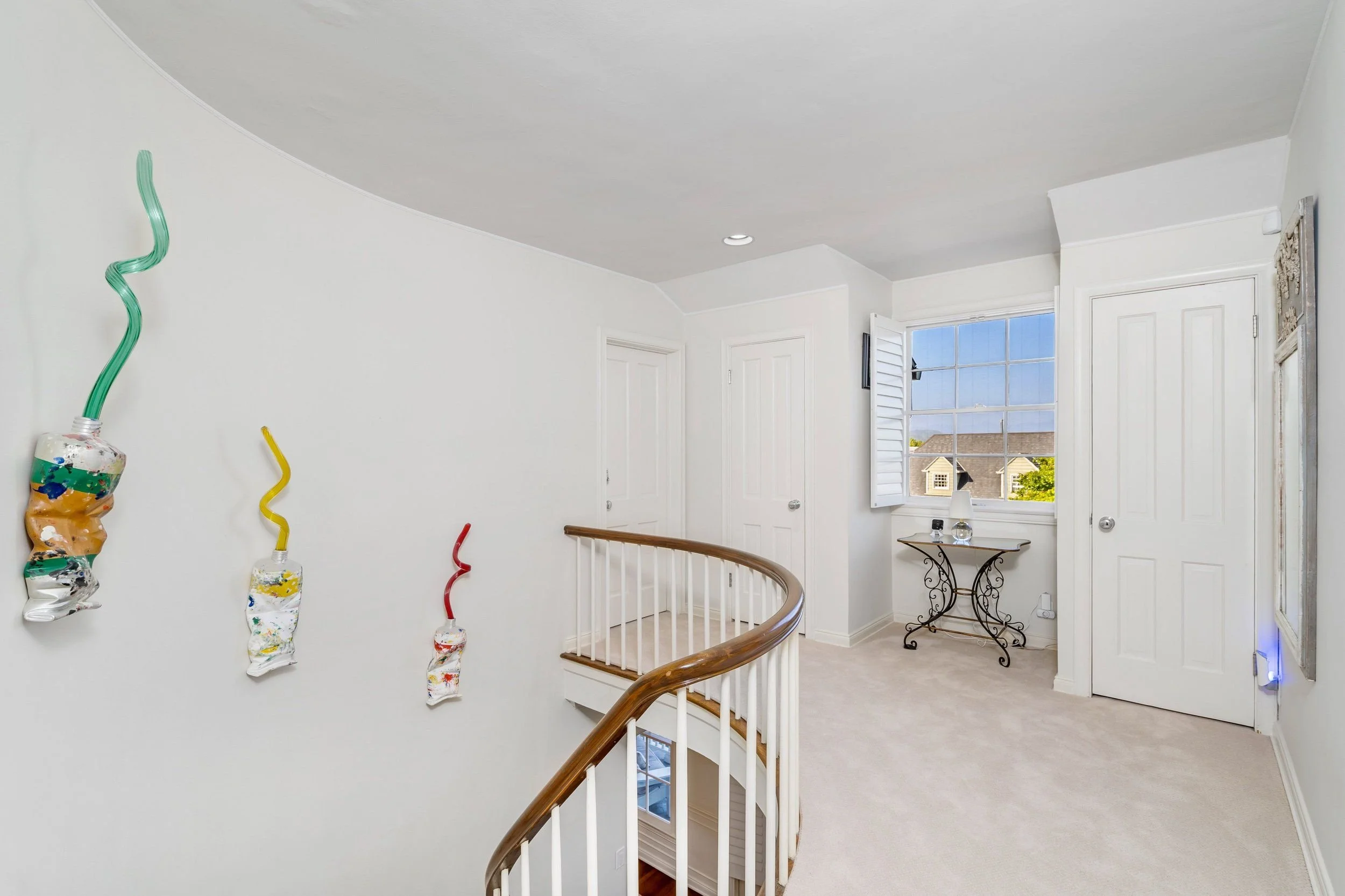 Interior of a house with white walls and ceiling, featuring decorative wall art and a window with a view of neighboring houses, with a staircase with a wooden handrail in the foreground.