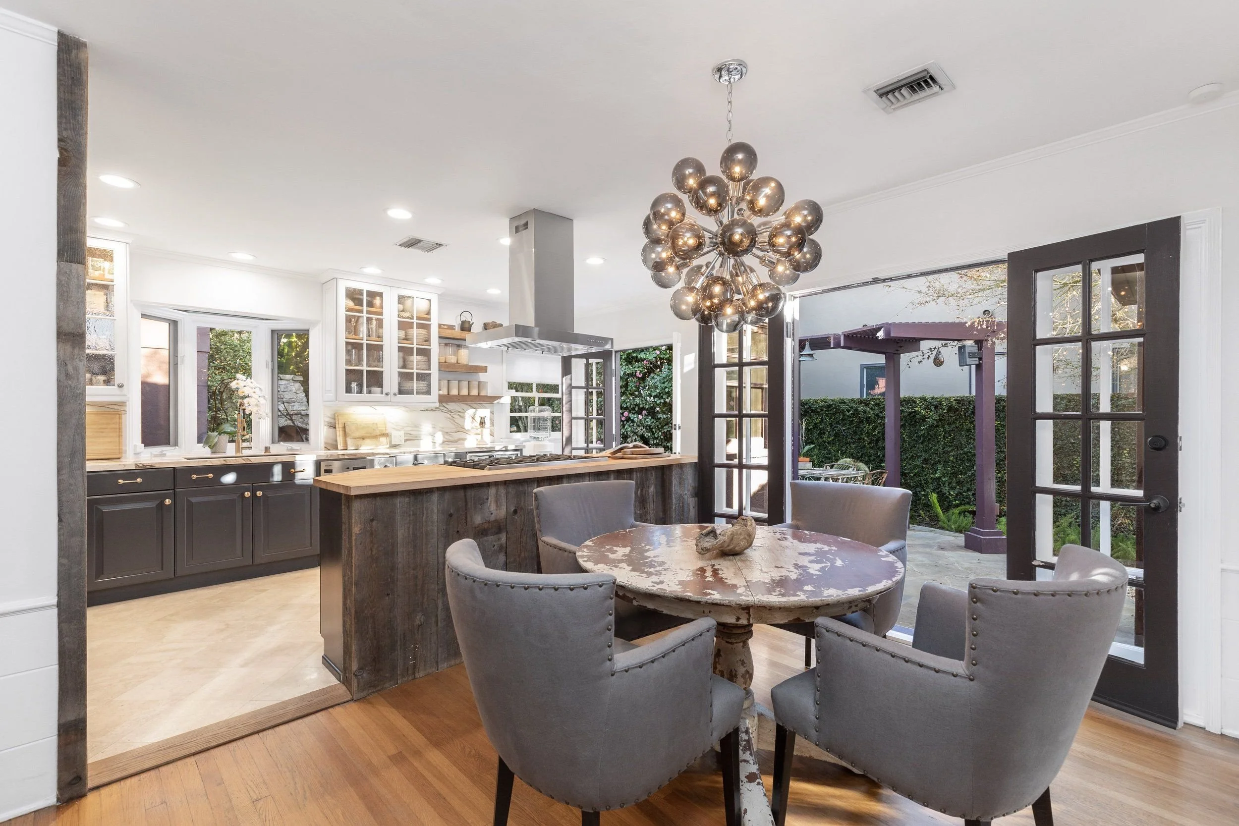 View of a cozy dining area attached to a kitchen, with a round wooden table surrounded by tufted gray chairs, a modern chandelier, and a large glass door opening to a backyard outdoor space with greenery.