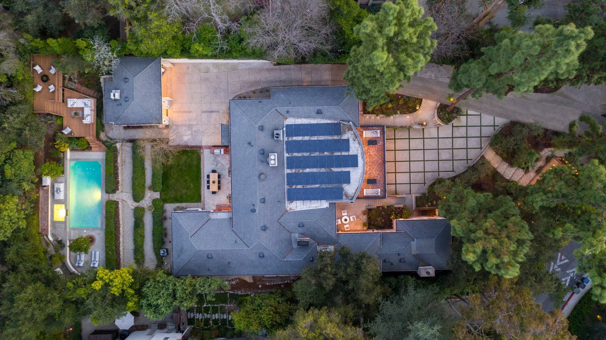 An aerial view of a residential backyard showing a swimming pool, a patio with outdoor furniture, a house with solar panels on the roof, a driveway, and surrounding greenery including trees and shrubs.