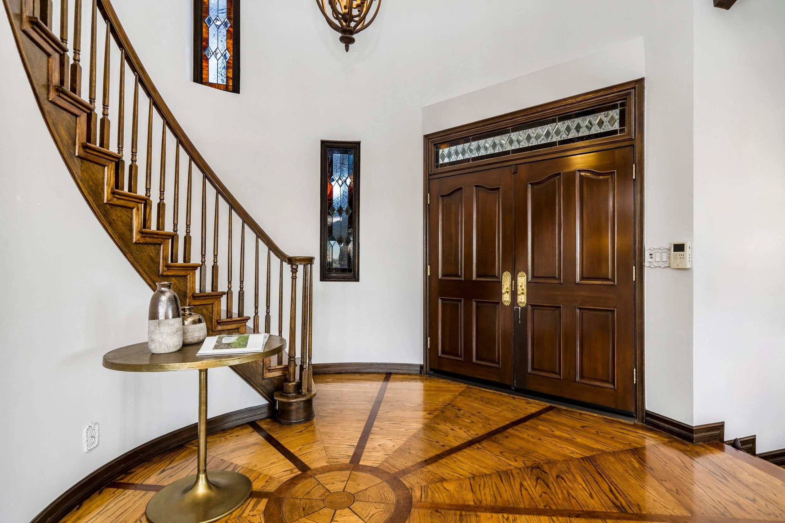 Entryway with hardwood flooring, a wooden front door with brass handles, stained glass windows above and beside the door, a staircase with wooden railing, a round side table with decorative vases, and a wall-mounted control panel.