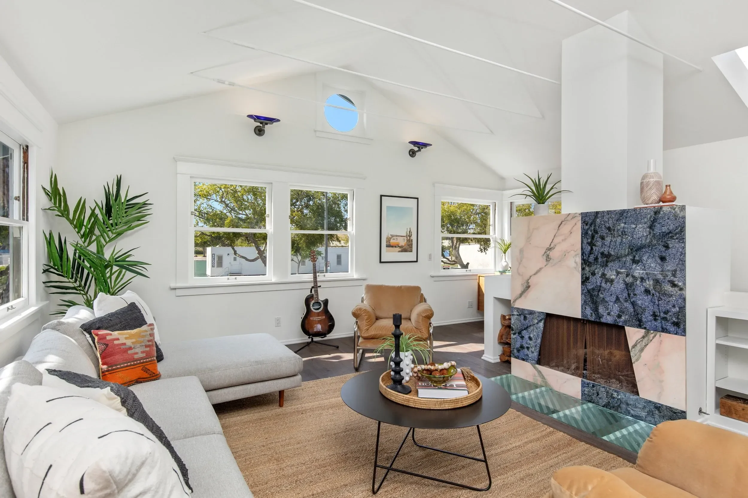 Living room with white walls, large windows, a beige sofa with decorative pillows, a guitar, indoor plants, a round black coffee table, and a fireplace with a marble facade.
