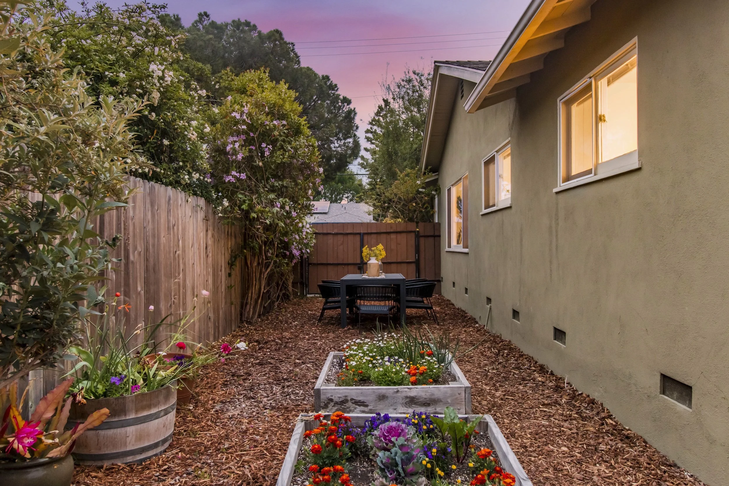 Backyard garden with wooden raised flower beds filled with colorful flowers, a table with chairs, and a house wall with windows at sunset.