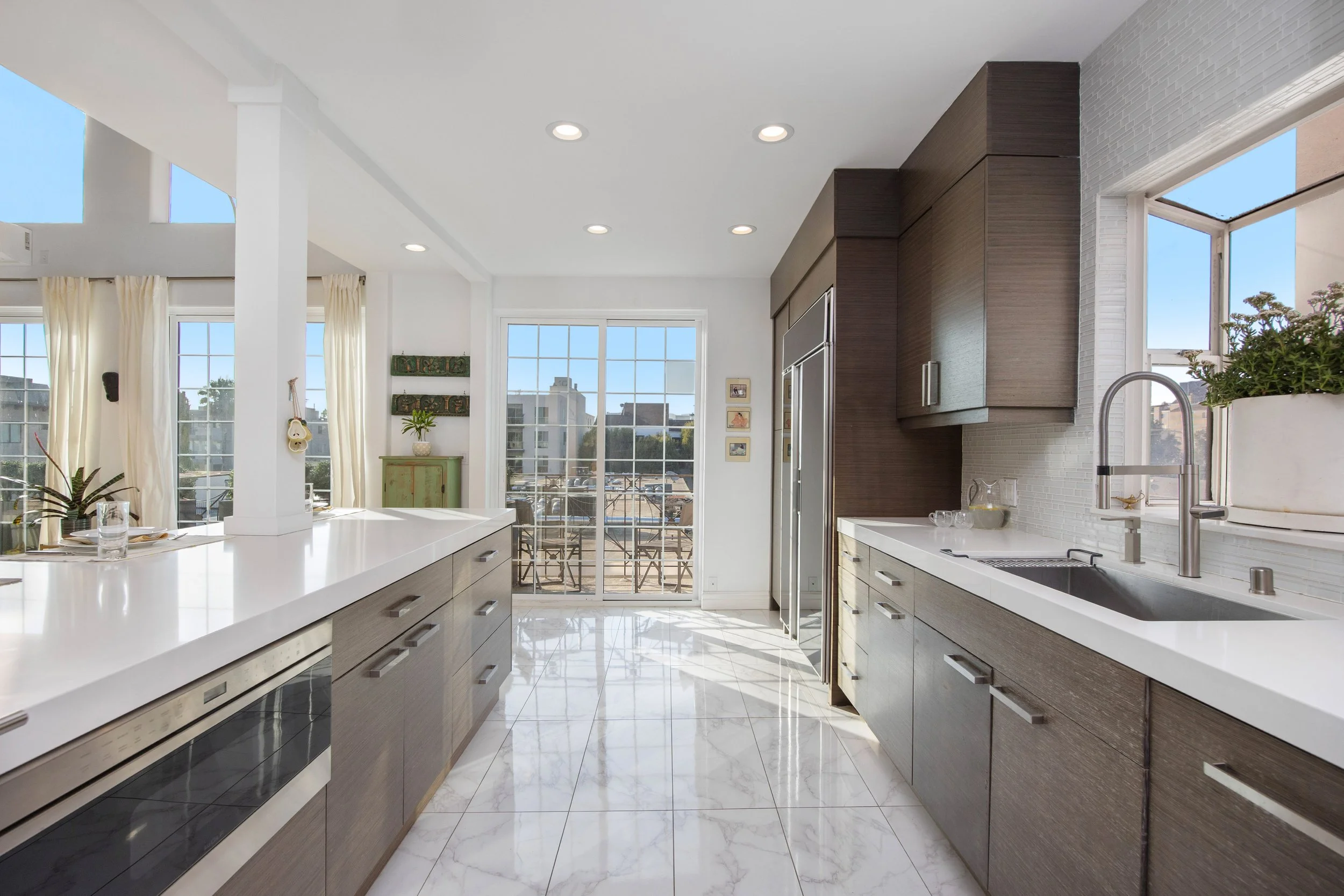 Modern kitchen with white countertops, wooden cabinets, large windows, and a view of a balcony.
