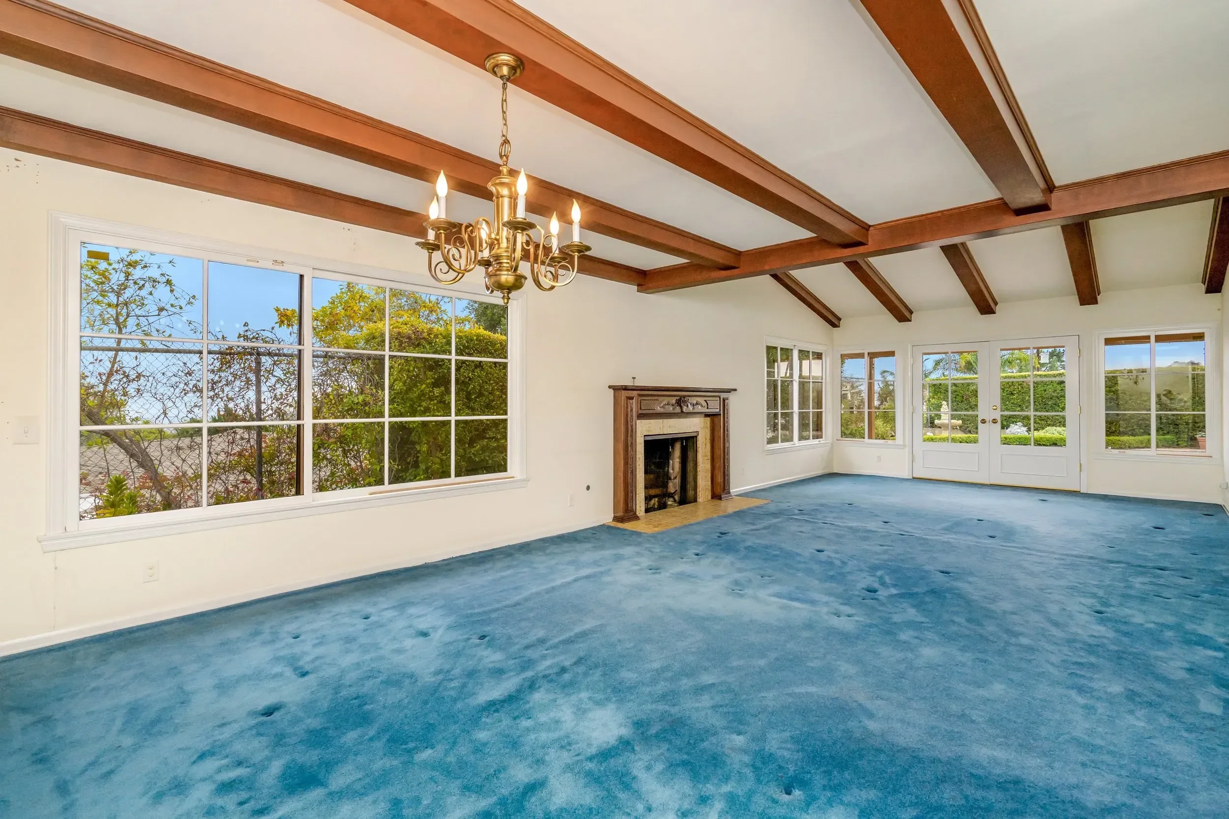 Empty living room with large windows, wooden ceiling beams, chandelier, fireplace, blue carpet, and view of trees outside.