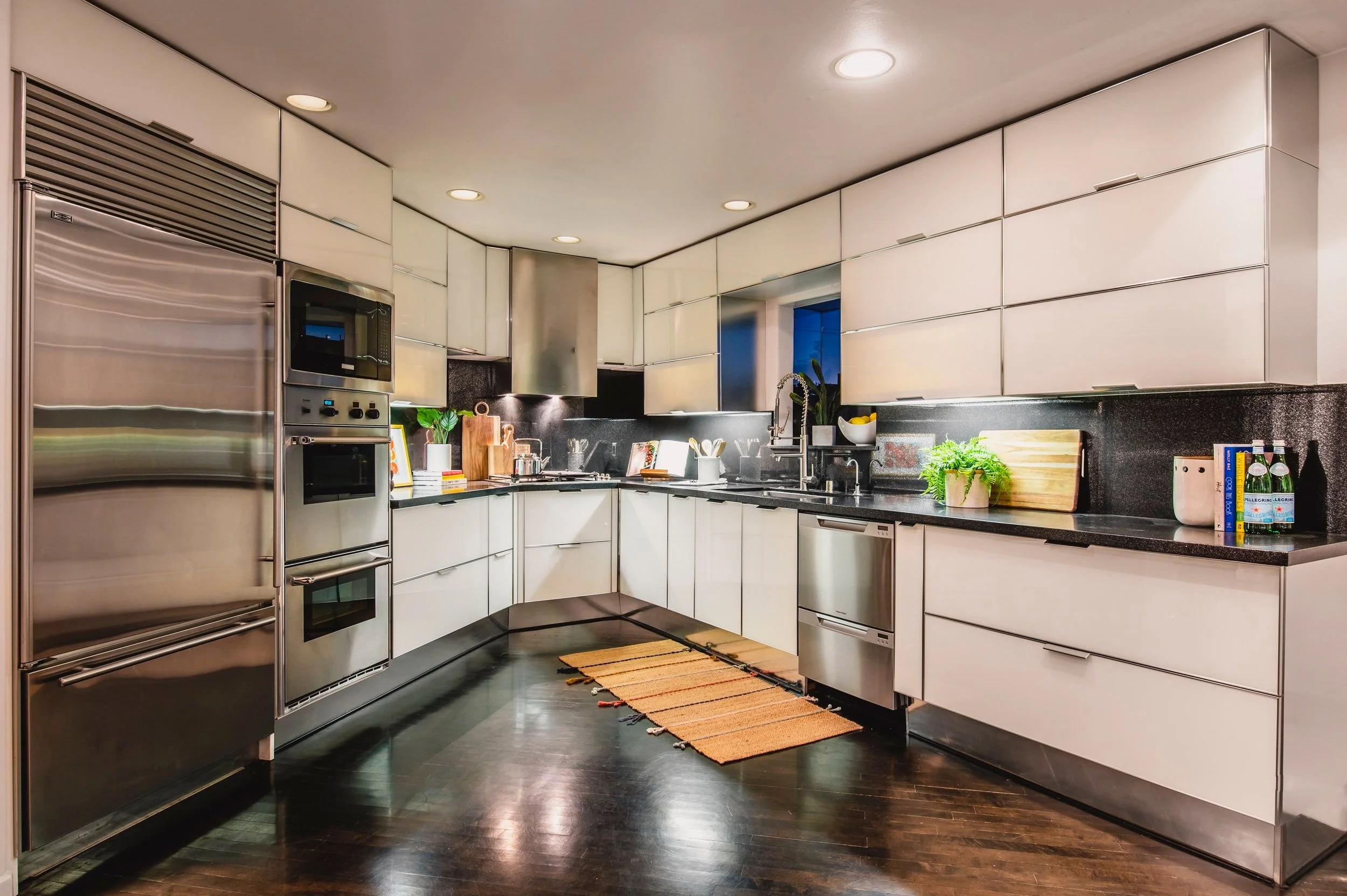 Modern kitchen with white cabinets, black countertops, stainless steel appliances including refrigerator, oven, and microwave, and a black backsplash. There is a sink with a faucet, various plants, books, and kitchen utensils on the counter, and a sm
