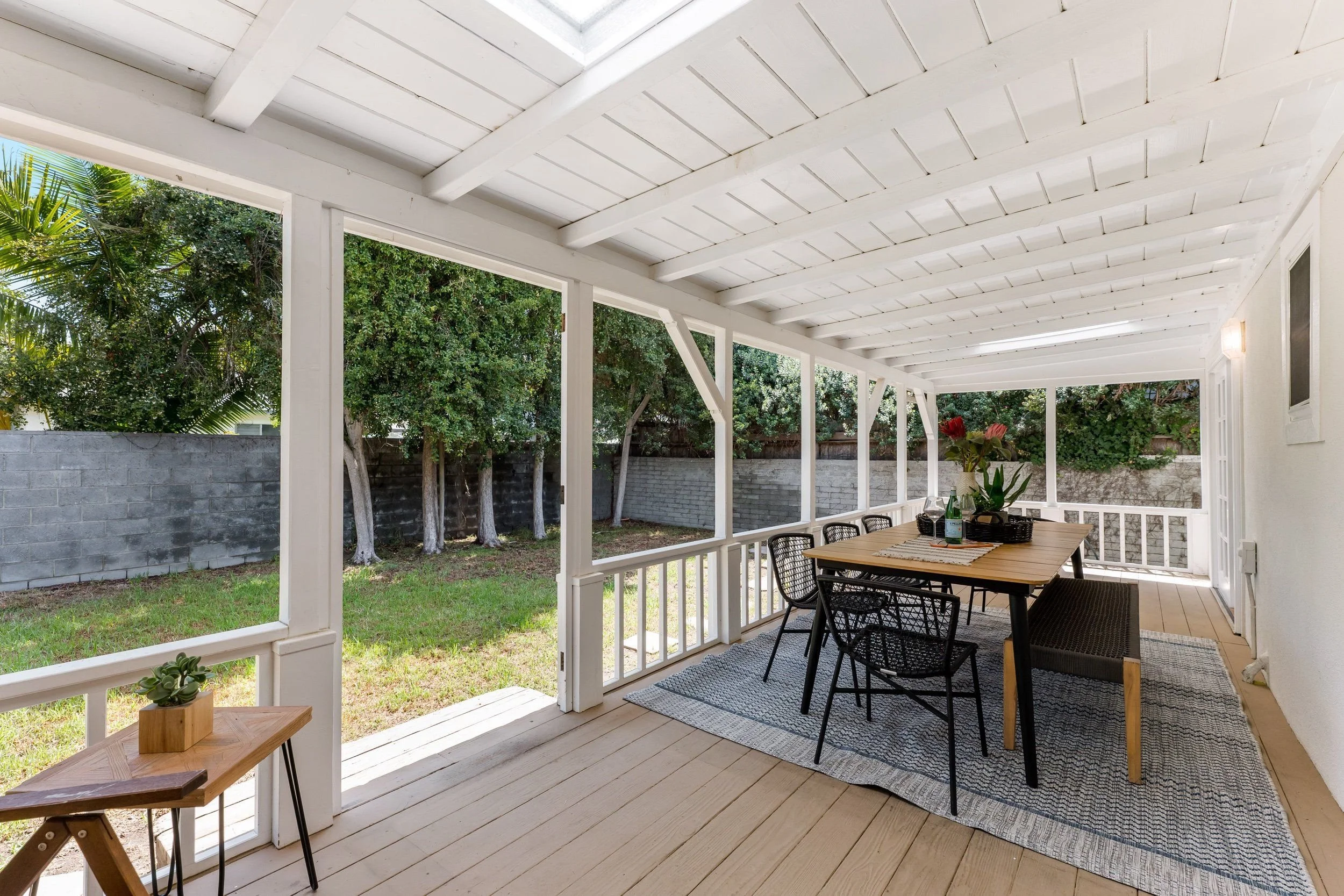 A covered porch with a wooden dining table, black chairs, and a potted plant, surrounded by green trees and a gray concrete wall.