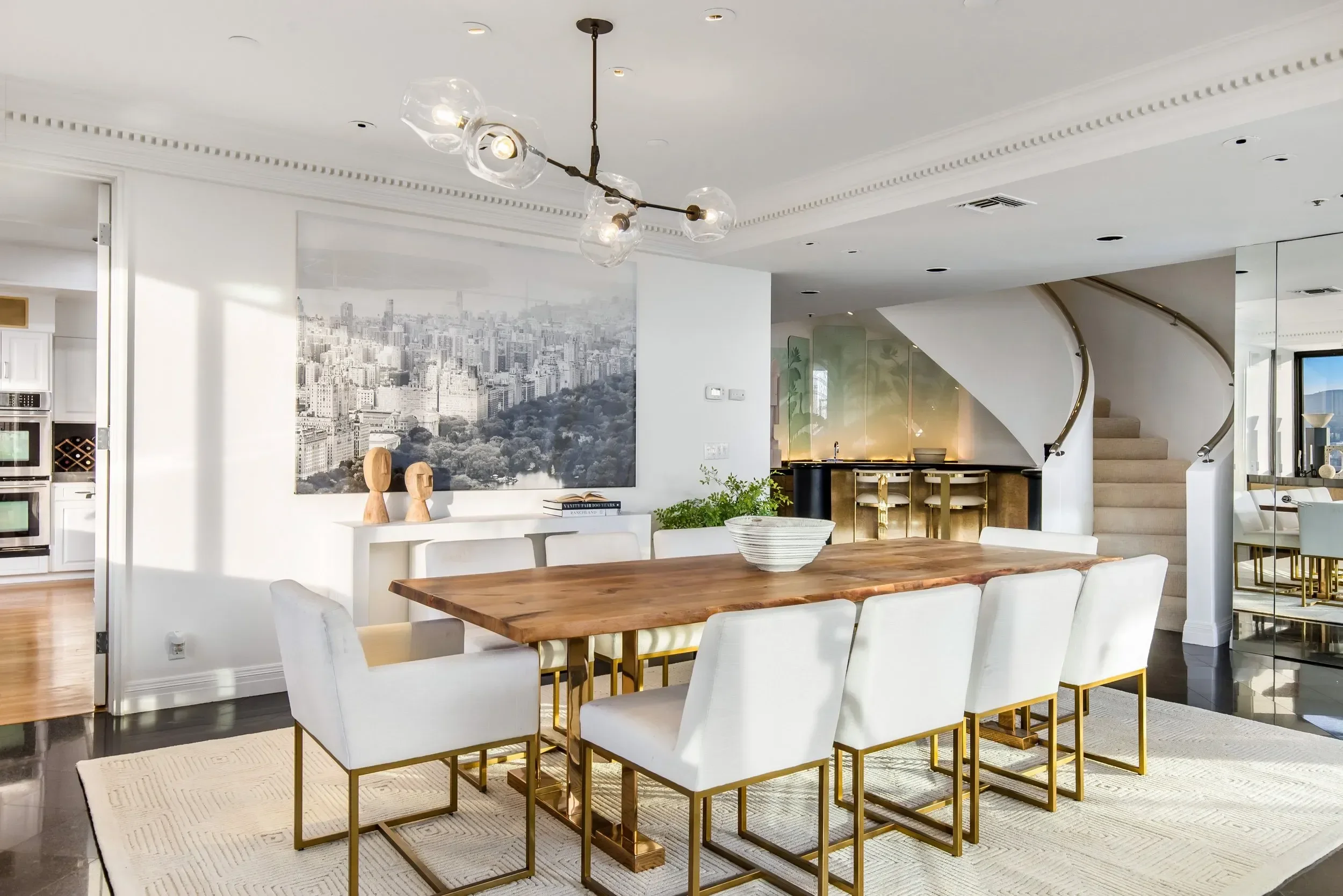 Modern dining room with a large wooden table surrounded by white chairs with gold legs. A large black and white cityscape wall art is on the wall, and a unique chandelier with glass bulbs hangs from the ceiling. The room features white walls, crown m