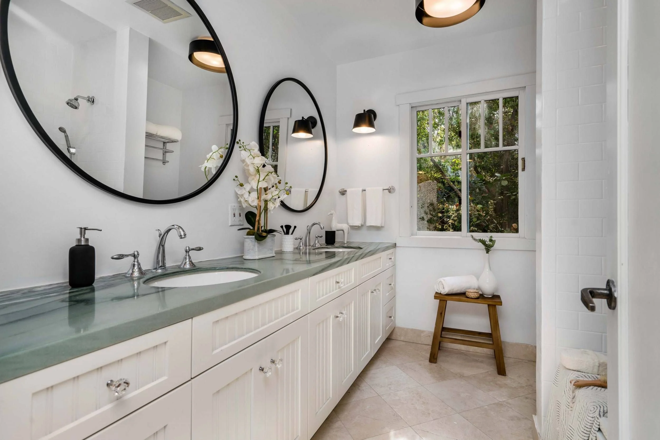 A modern bathroom with twin oval mirrors above a white vanity countertop with two sinks. Decor includes a potted orchid, black soap dispenser, and toothbrush holder. Black wall sconces provide lighting, and a large window shows greenery outside. Othe