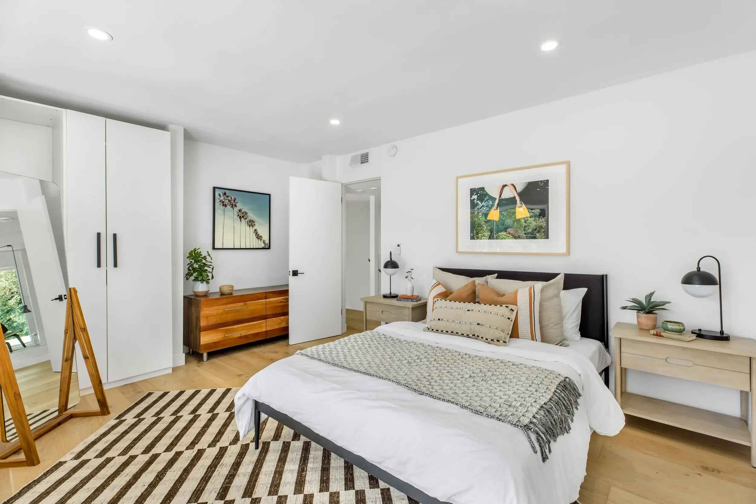 Modern bedroom with white walls, bed with striped bedding and multiple pillows, wooden nightstands with lamps, framed artwork on the wall, and a rug with black and white geometric pattern.
