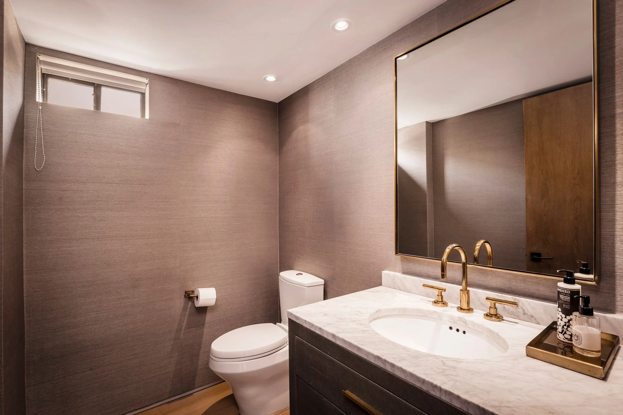 Modern bathroom with beige textured walls, a white toilet, and a marble countertop with a gold faucet. A large mirror with a gold frame is above the sink. Small bottles of soap and lotion are on a tray by the sink. There is a small high window with b