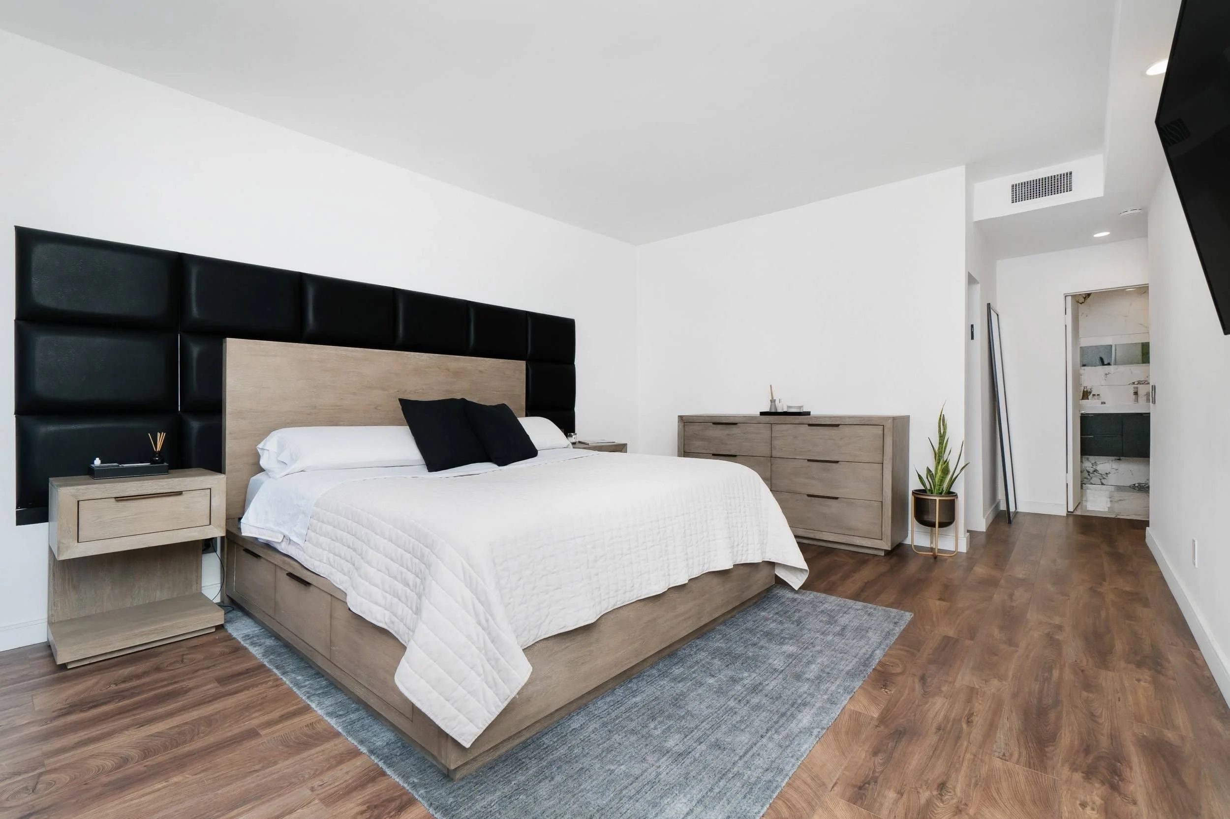 Modern bedroom with a wooden bed frame, white bedding, black pillows, wooden nightstands, a wooden dresser, a tall plant, and a flat-screen TV mounted on the wall.