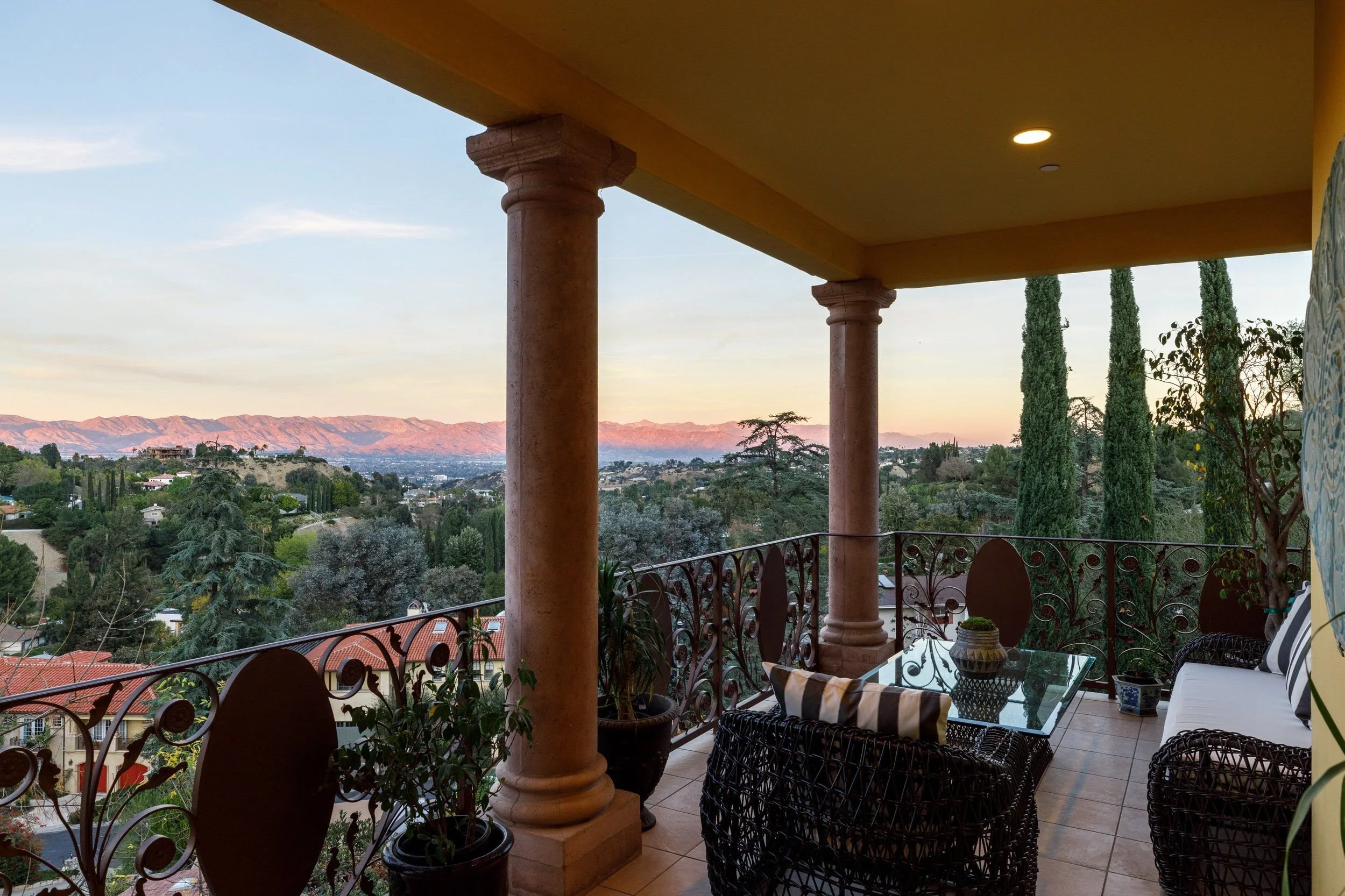 Balcony with decorative iron railing, potted plants, and outdoor furniture overlooking a landscape of trees, houses, and mountains at sunset.