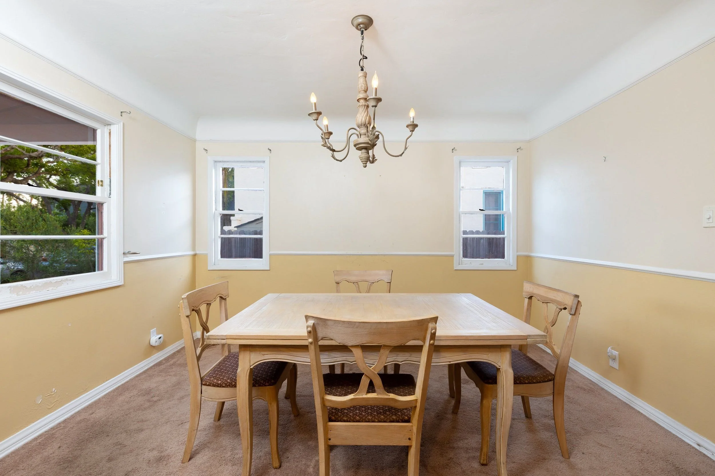 Dining room with square wooden table and six matching chairs, three windows with white frames on the beige walls, a chandelier with candle-shaped lights hanging from the ceiling, and a brown carpeted floor.