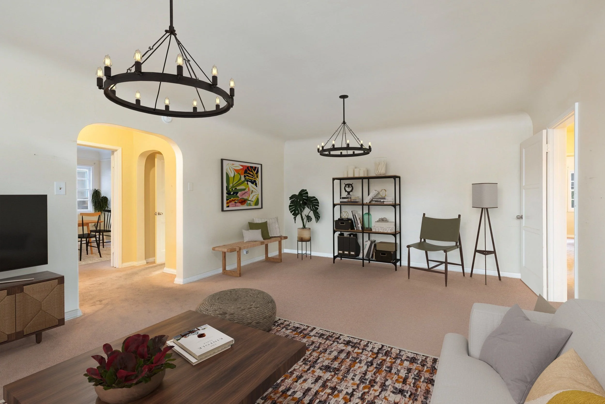 Living room with two black circular chandeliers, a light-colored sofa with pillows, a wooden coffee table with books and a plant, a patterned rug, and a black metal bookshelf with decorative items. Yellow arched doorway leads to a dining area with a 