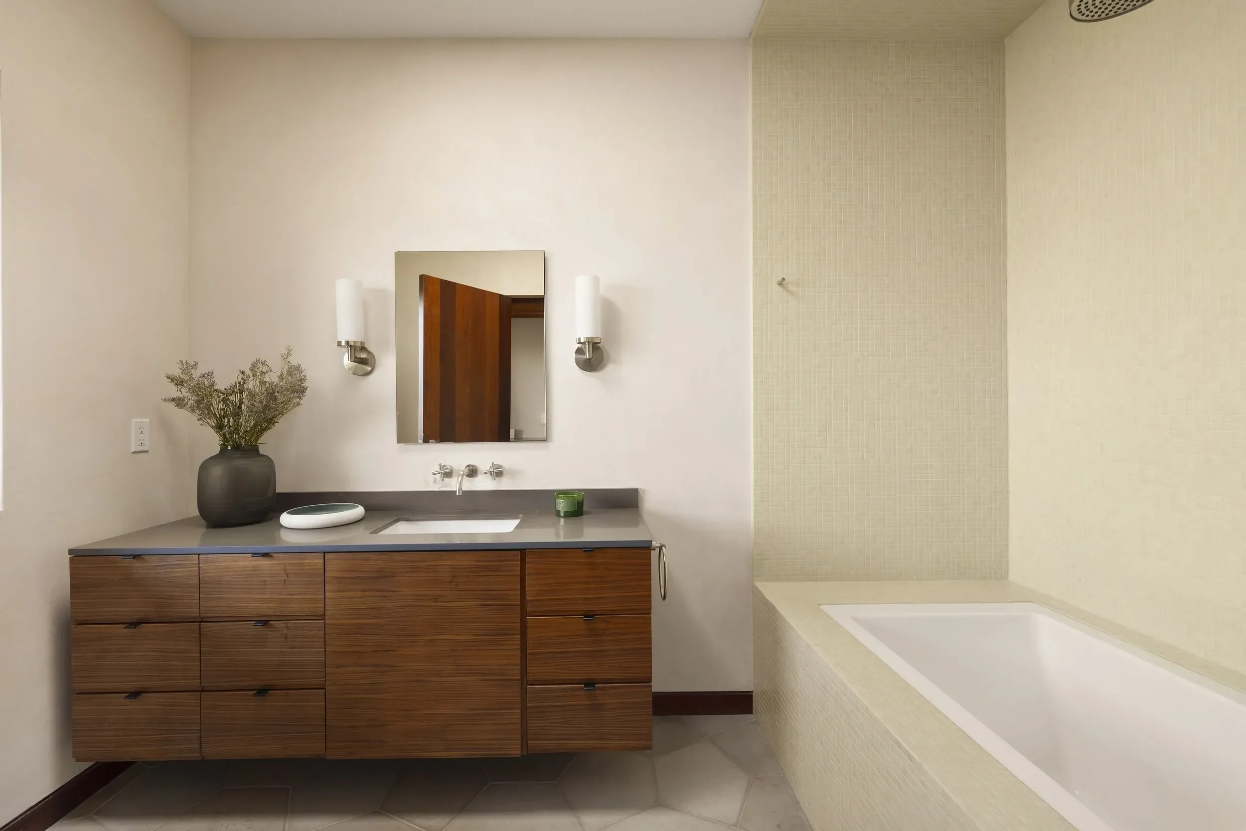 Modern bathroom with a wooden vanity, rectangular mirror, white sink, black countertop, decorative plant, wall-mounted light fixtures, and a walk-in shower with beige tiles.