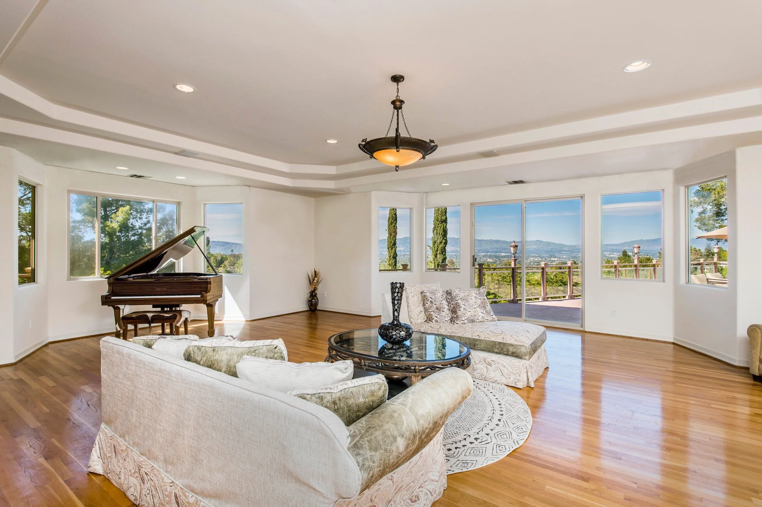 Spacious living room with large windows showing a scenic view, hardwood floors, a white sofa and loveseat with patterned cushions, a glass coffee table with decorative vases, a grand piano, and a chandelier hanging from the ceiling.