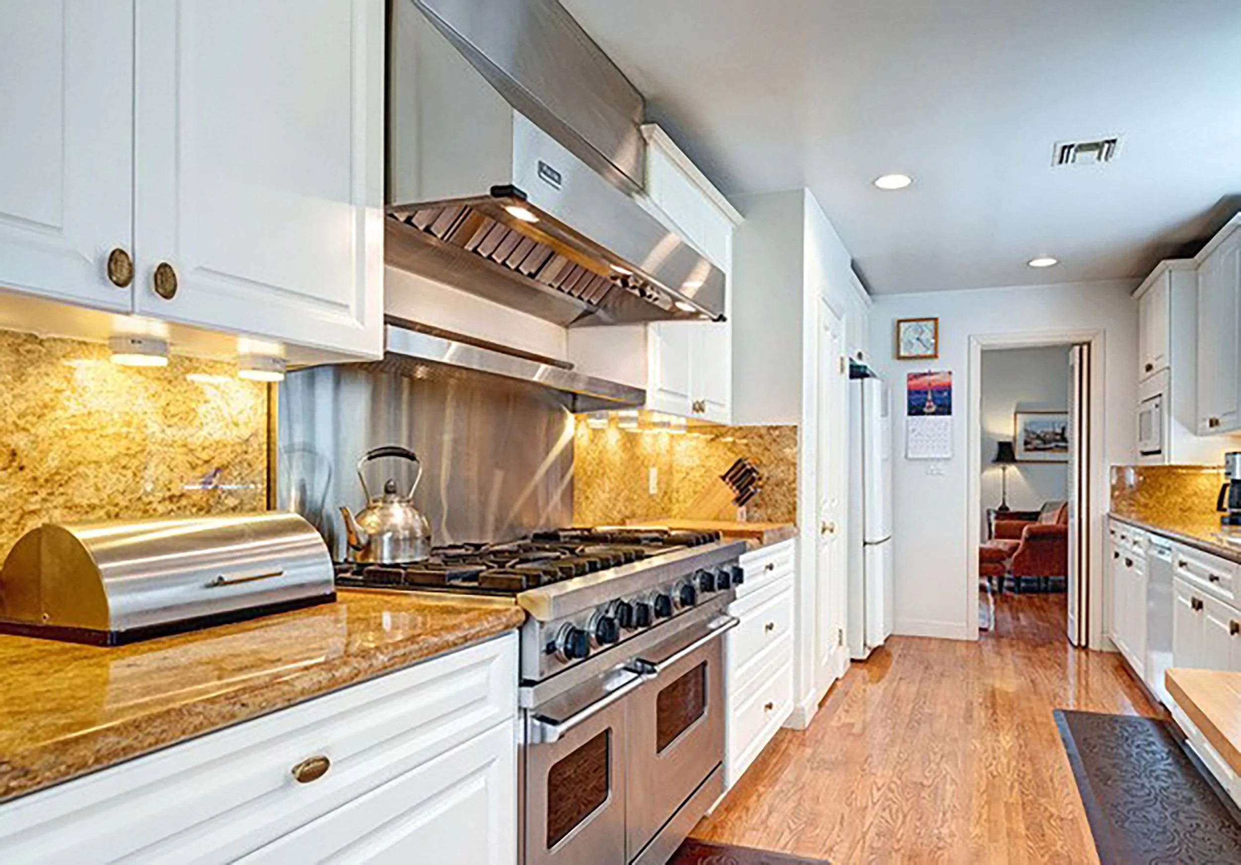 A kitchen with white cabinets, a gas stove with six burners, and a granite countertop. There is a stainless steel range hood above the stove, and a roll of paper towels and a kettle on the counter. In the background, a living area with wall art, a ca