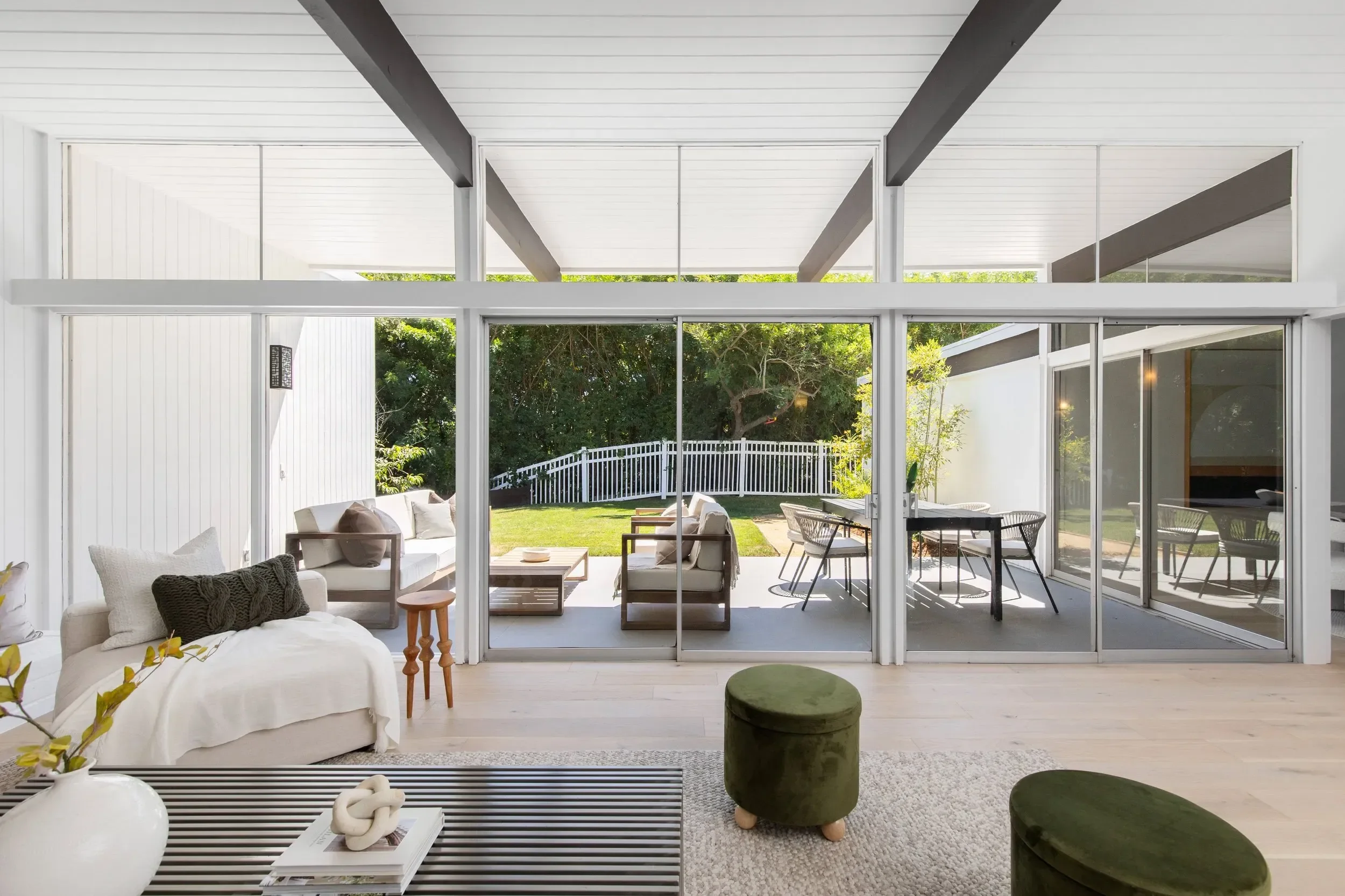 Living room with white walls and ceiling, large sliding glass doors opening to backyard patio with outdoor seating and white picket fence, and indoor furniture including a white couch, green ottomans, and a black and white striped coffee table.
