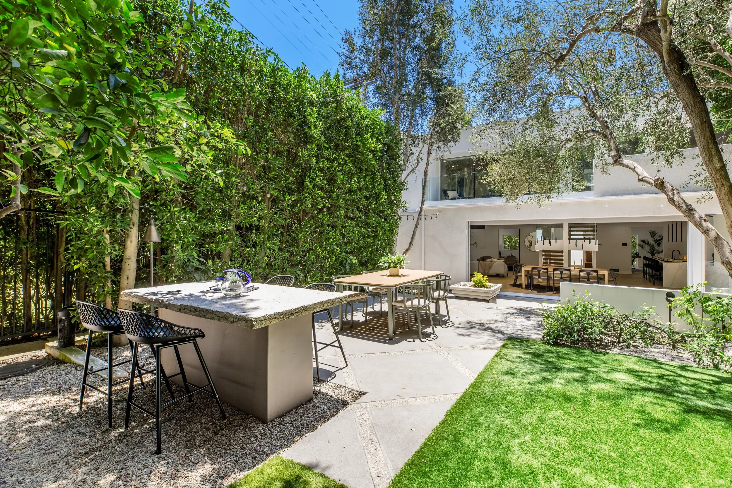 A modern backyard patio area with outdoor dining table, chairs, and lush green trees surrounding the space, with views of a contemporary white house.