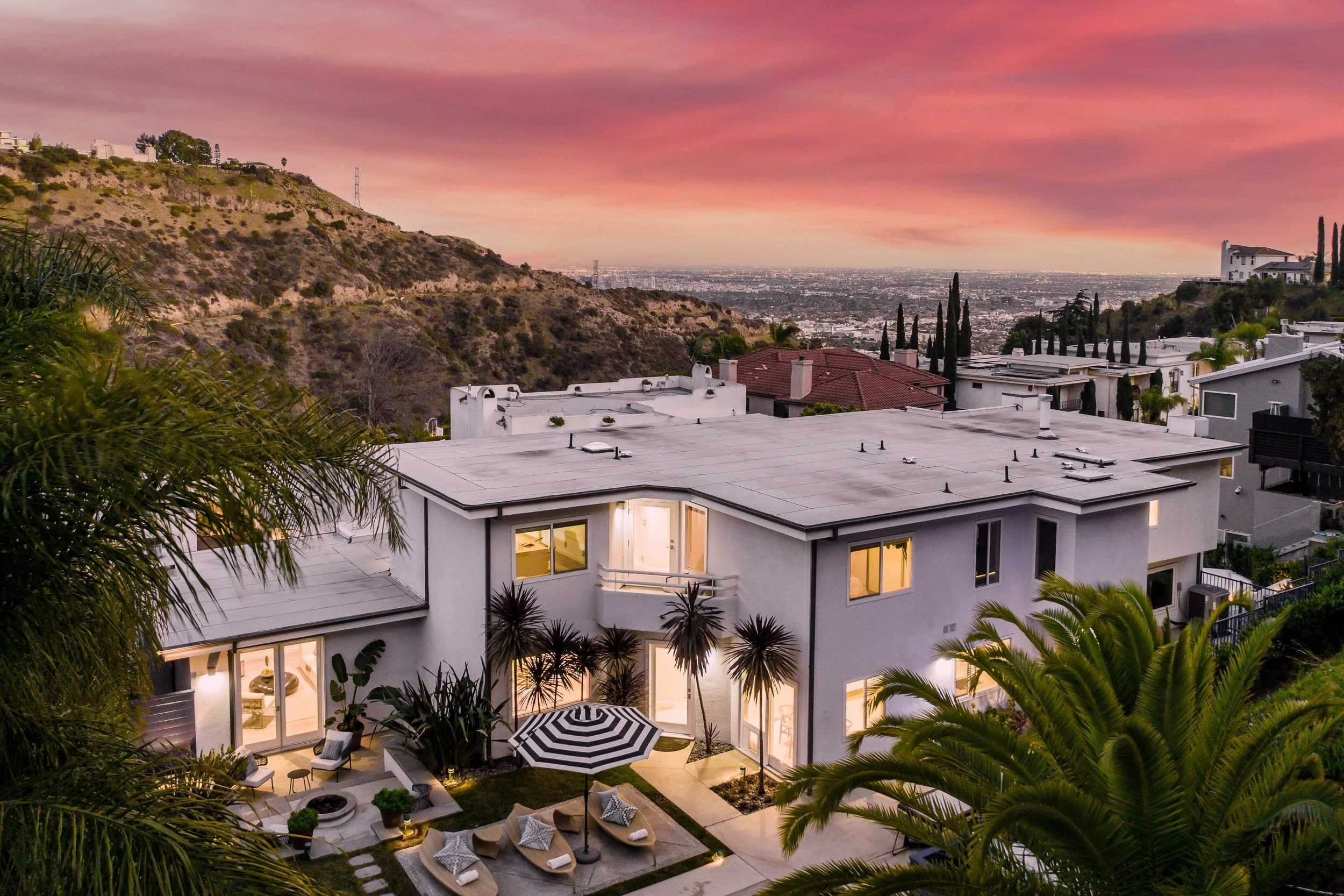 A modern white house with large windows, surrounded by palm trees and outdoor seating, overlooking a hilly landscape and city skyline at sunset with pink and purple sky.