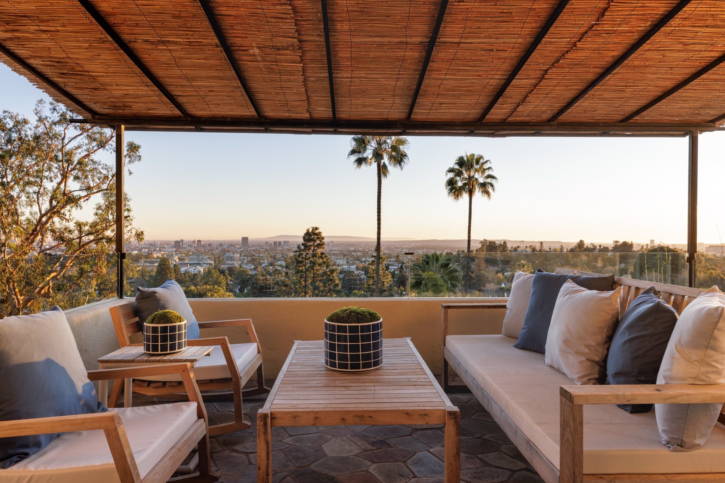 Outdoor seating area with two sofas and wooden tables, overlooking a city skyline with palm trees during sunset.