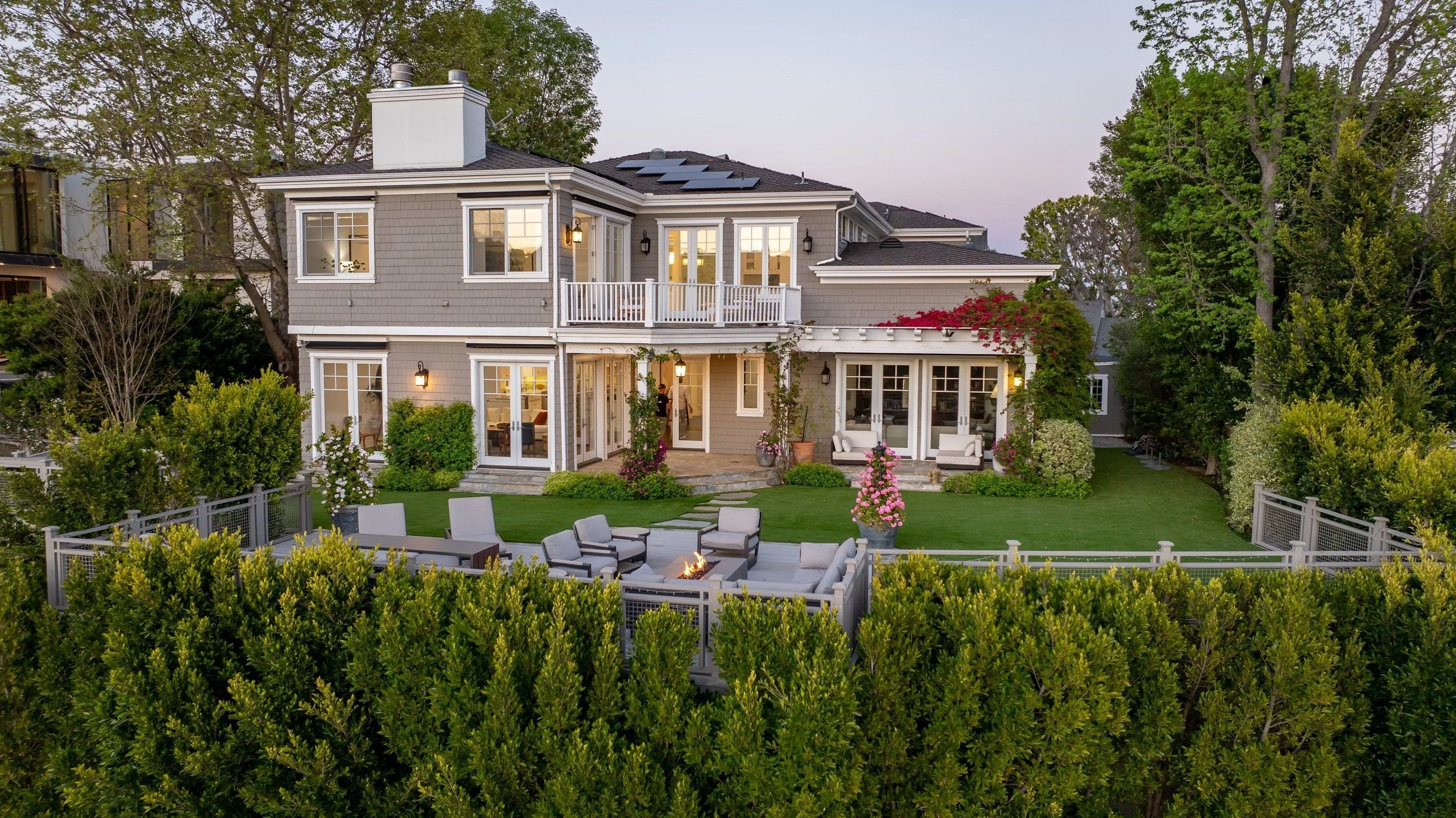 Two-story gray house with large windows and a backyard patio with seating, surrounded by green trees and bushes.
