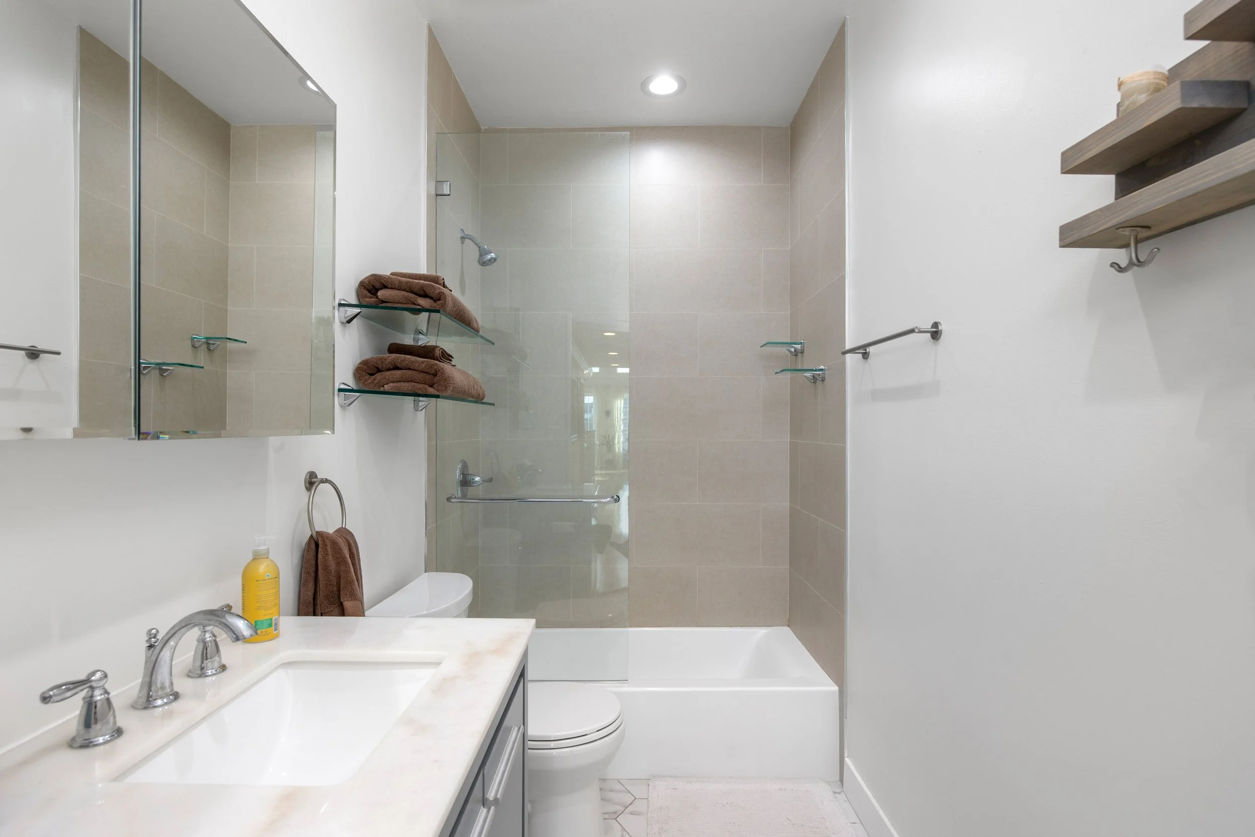 A modern bathroom with a white sink with silver fixtures, brown towels on glass shelves, a bottle of yellow soap, a toilet, a bathtub with a glass shower door, and beige tiled walls.