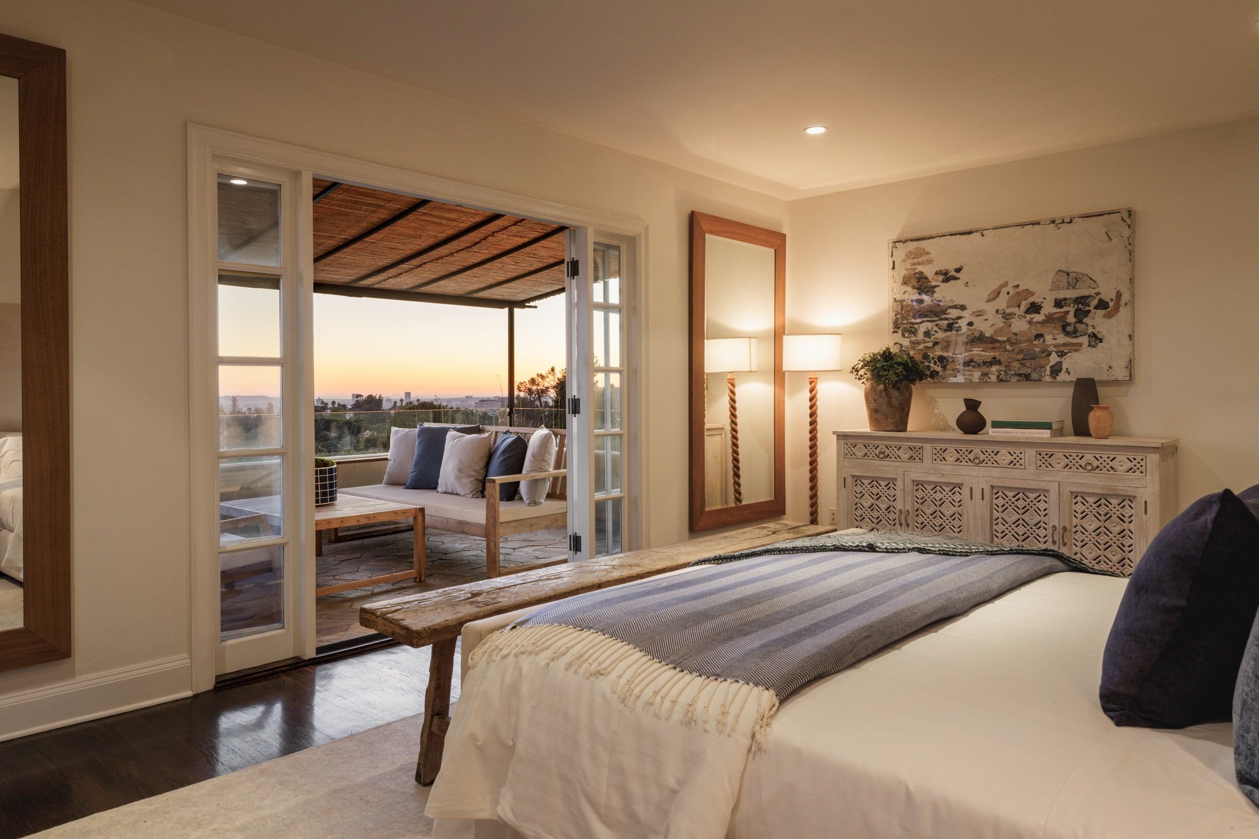 Room with a bed, a wooden bench, and French doors opening to a balcony with outdoor seating and a sunset view.