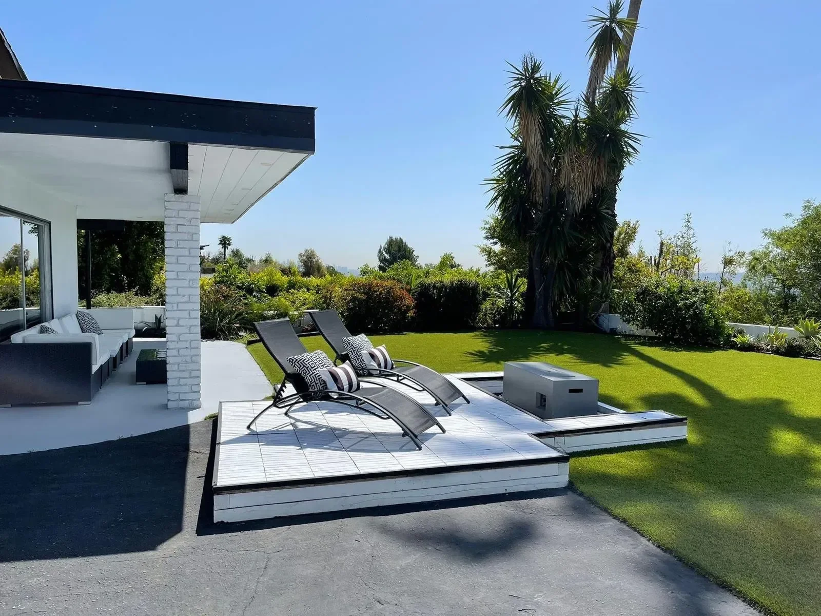 Modern outdoor patio with lounge chairs, a green lawn, and lush bushes and trees under a clear blue sky.