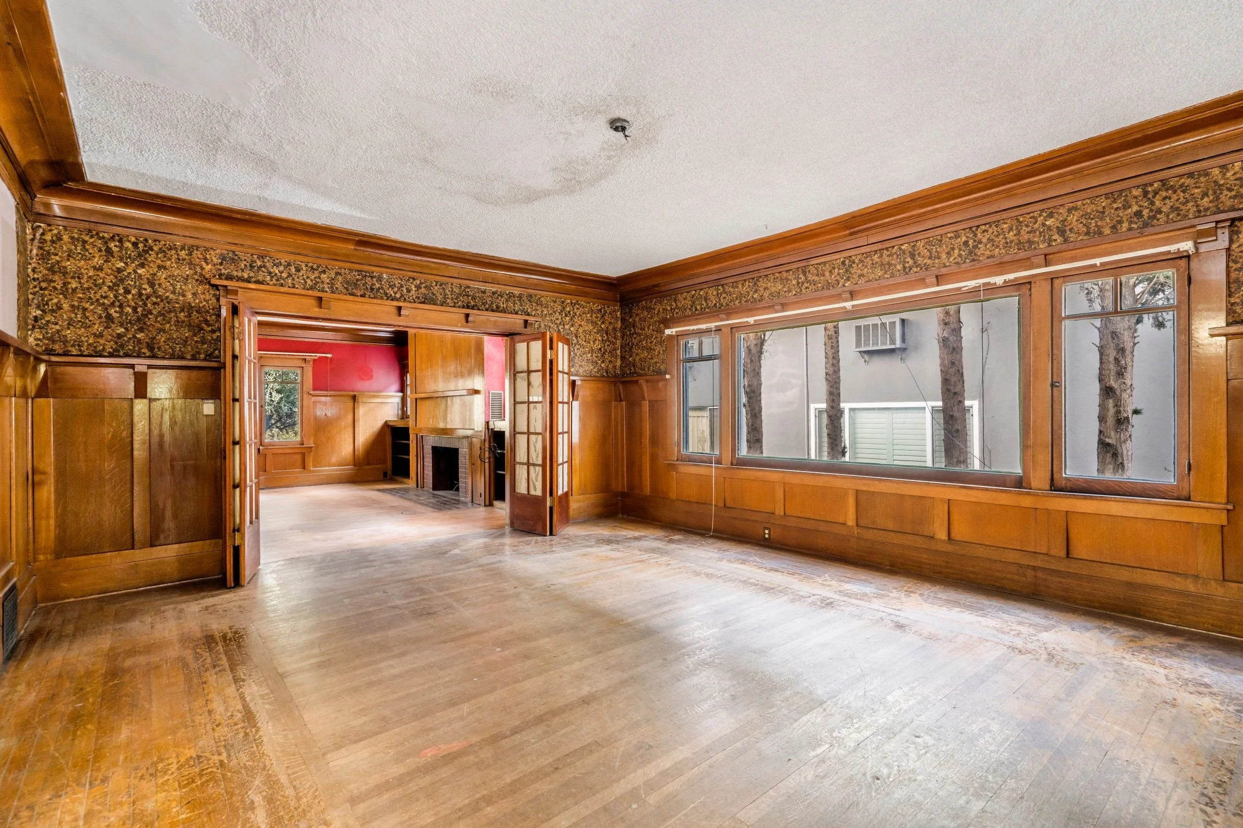 Empty living room with hardwood floors, large front window, wood paneling, built-in wooden shelves, and a brick fireplace.