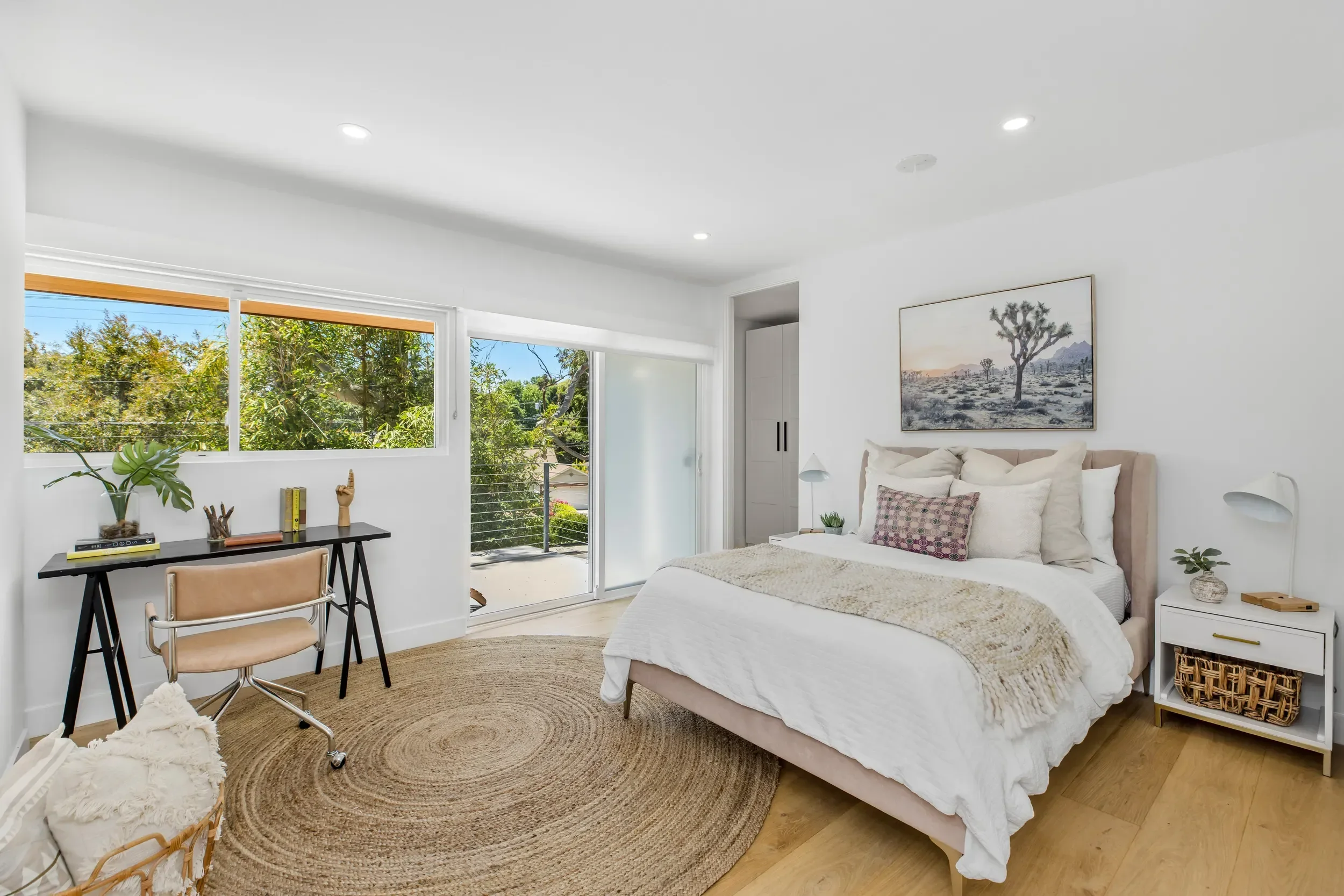 A bright, modern bedroom with a large bed with white bedding and decorative pillows, a nightstand with a plant, a framed desert landscape picture, a small desk with a chair and several books, and a sliding glass door leading to a balcony with greener