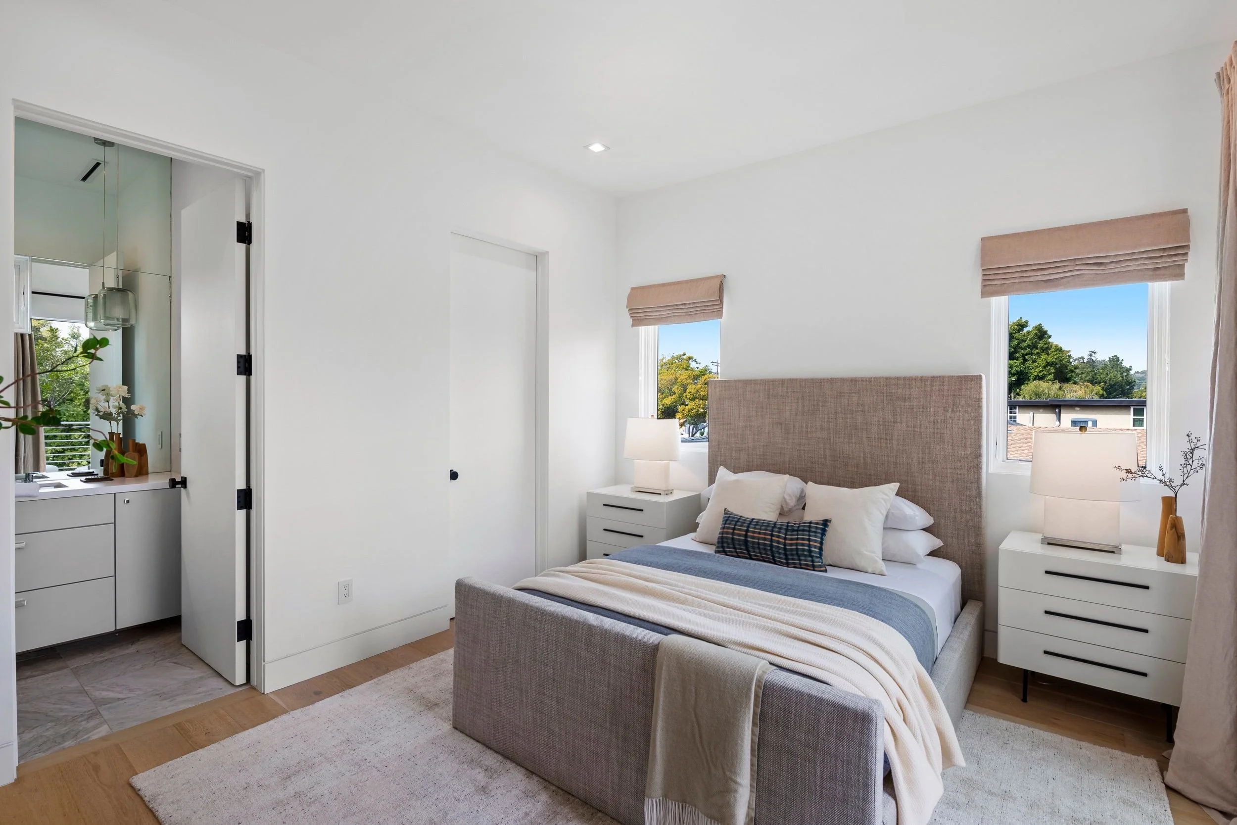 A modern bedroom with a beige upholstered bed, white nightstands, and cream-colored curtains on windows showing a view of trees outside.