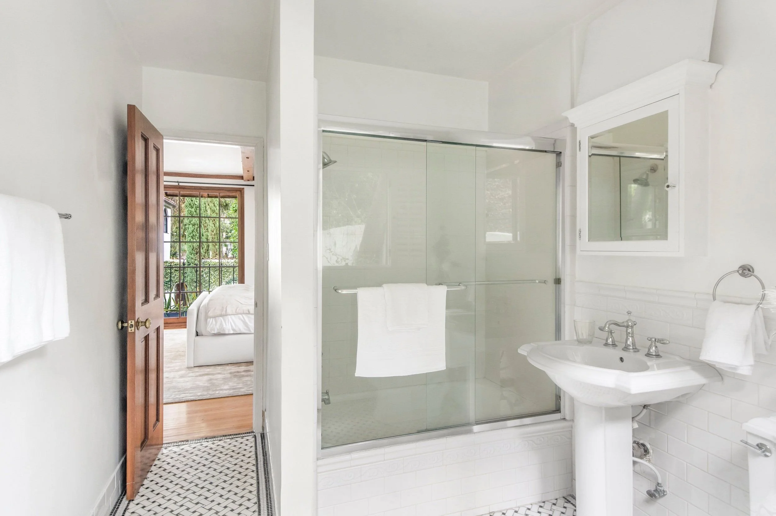 A white bathroom with a glass-enclosed shower, white sink with chrome fixtures, and a mirror. There is a towel hanging on a rail and a window showing trees outside.